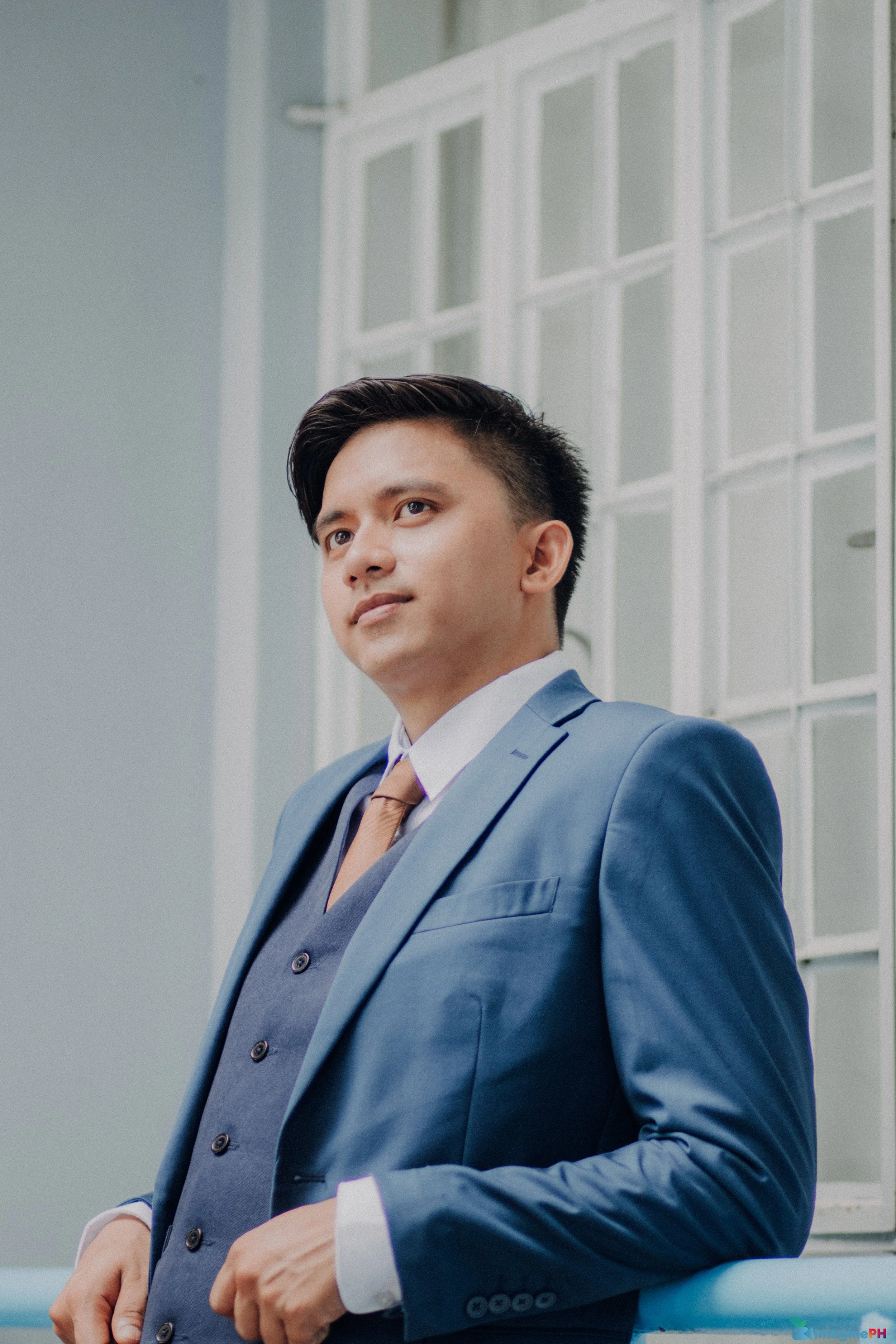 A young man in a blue suit and brown tie standing indoors near a large window.