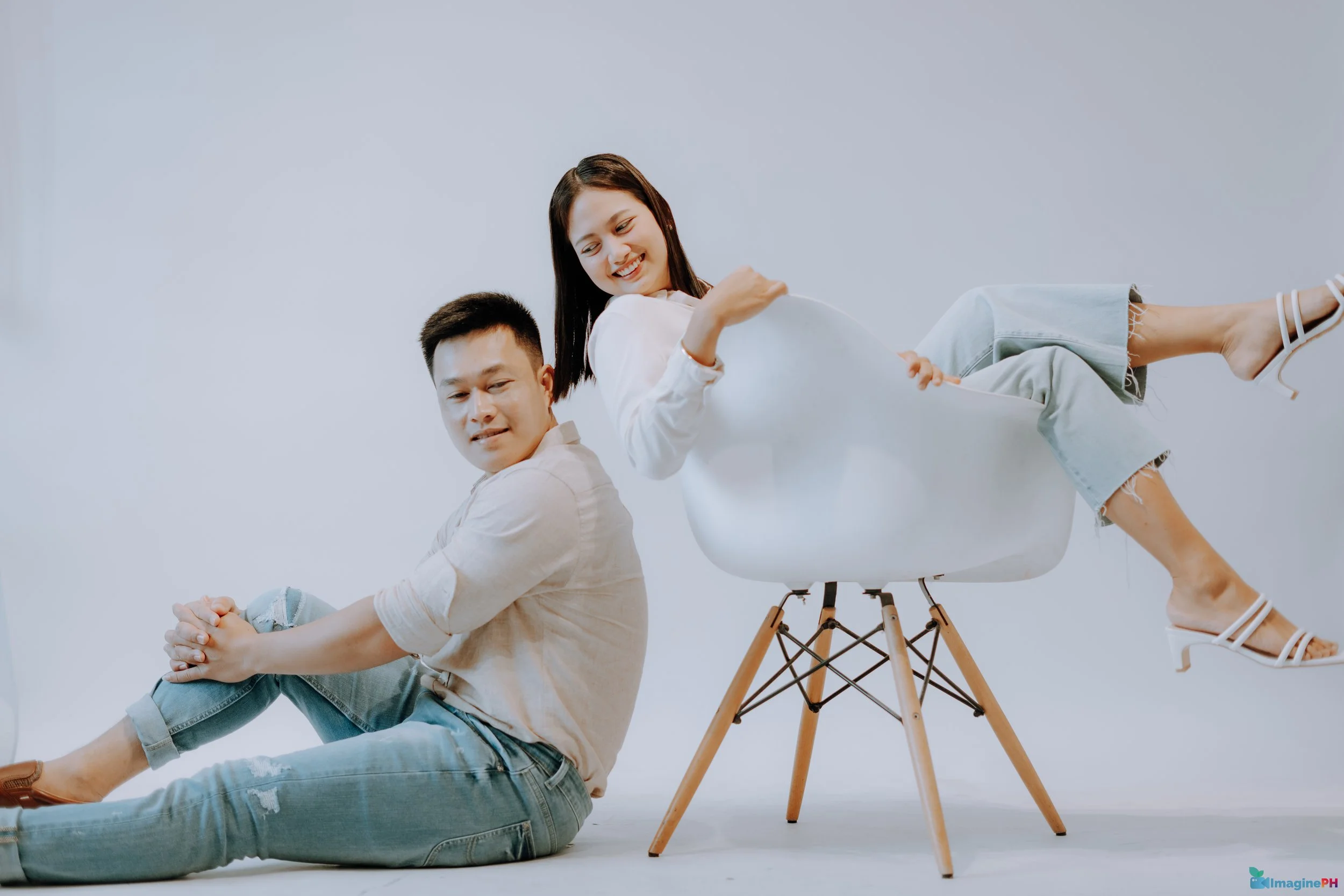 A young man sitting on the floor with his legs crossed and a young woman sitting in a white armchair, smiling and looking at each other against a plain white background.