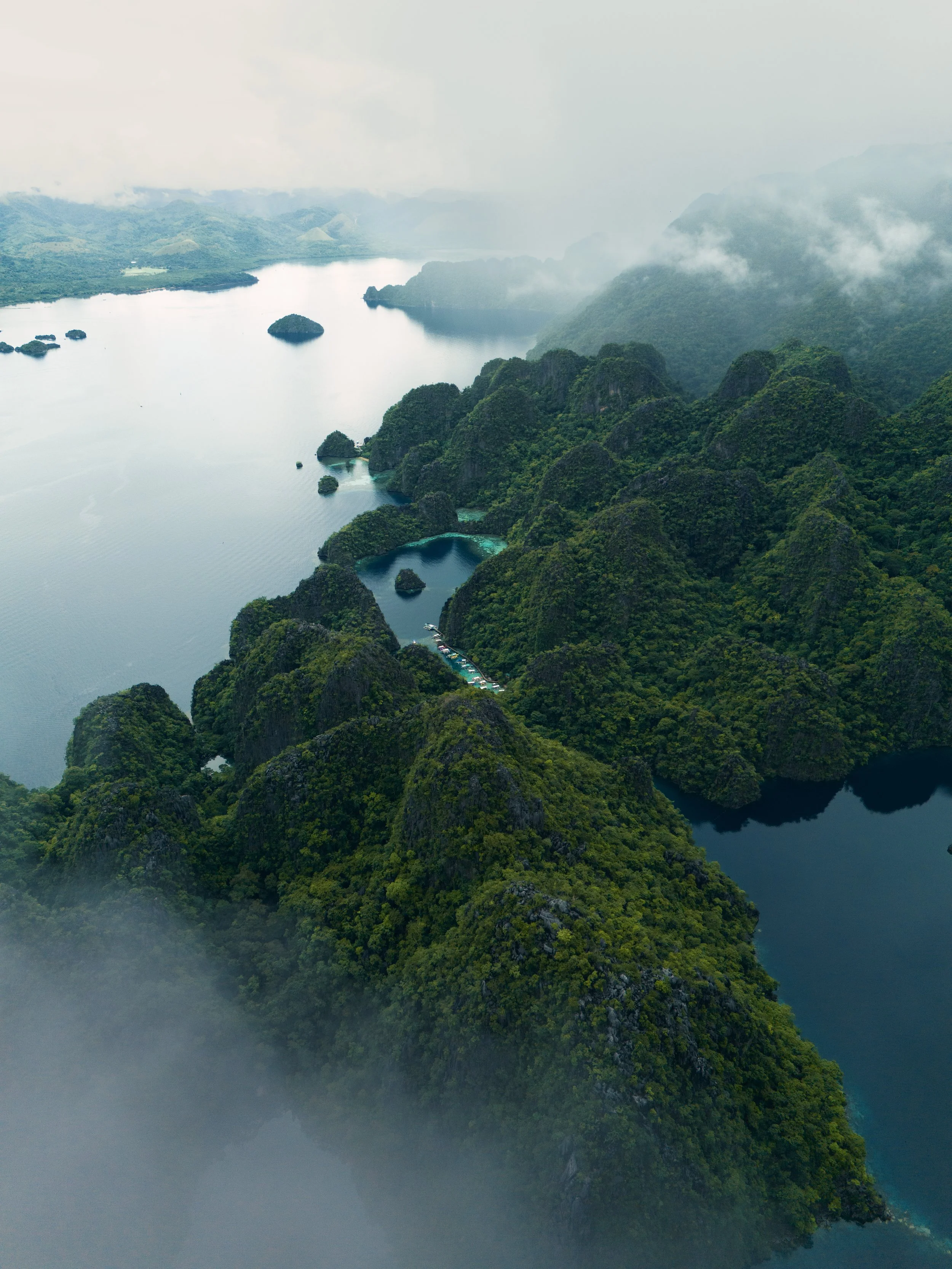 Aerial view of a lush green island with mountainous terrain, surrounded by calm river waters and smaller islands, with mountains shrouded in mist or clouds.