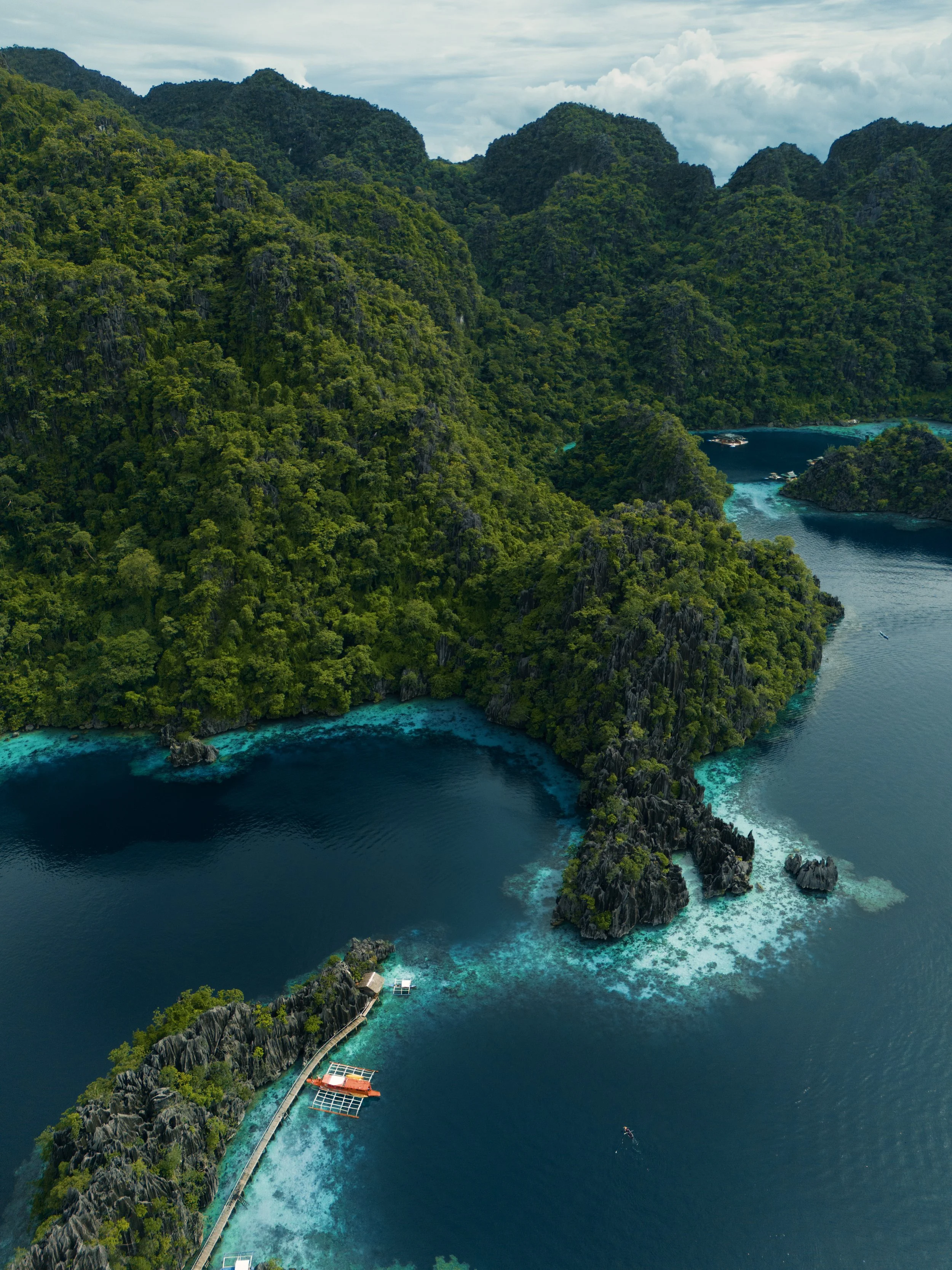 Aerial view of lush green islands with rocky cliffs surrounded by clear blue water, featuring a dock and some boats.