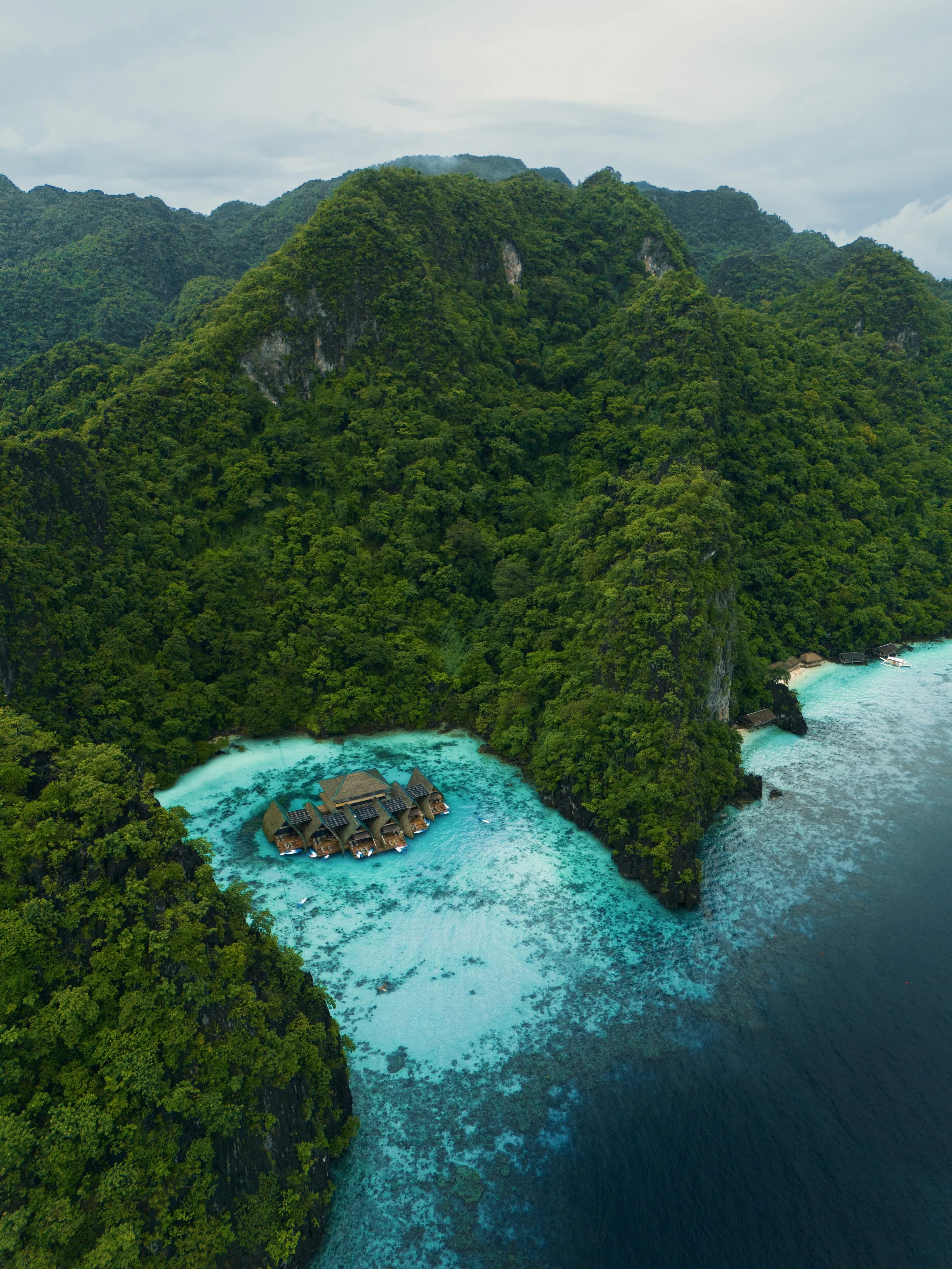 Overhead view of a tropical island with dense green forest, overwater bungalows, and turquoise ocean water.