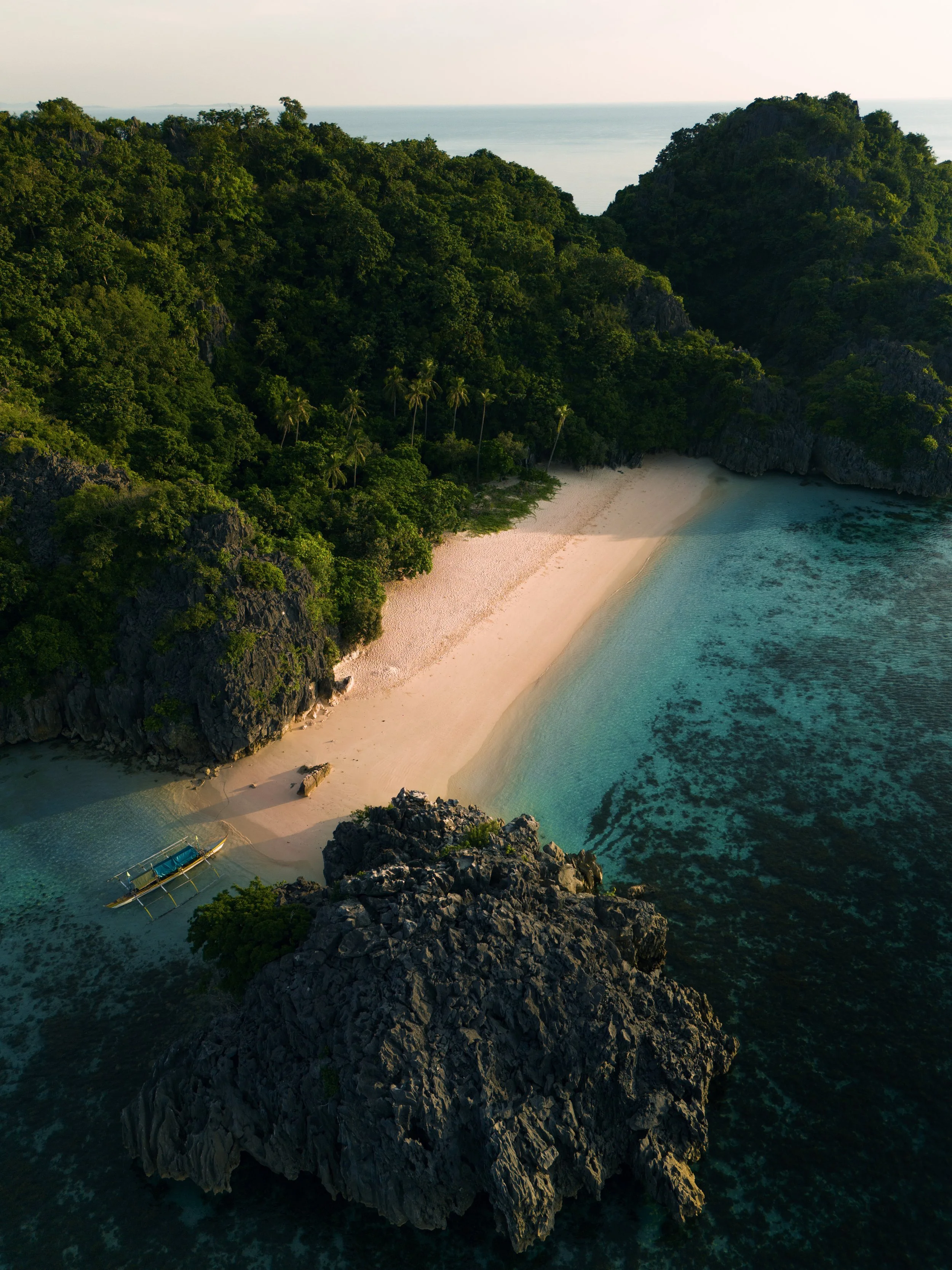 A secluded beach with white sand, surrounded by lush green trees and rocky cliffs, with clear turquoise water and a small boat anchored near the shore.