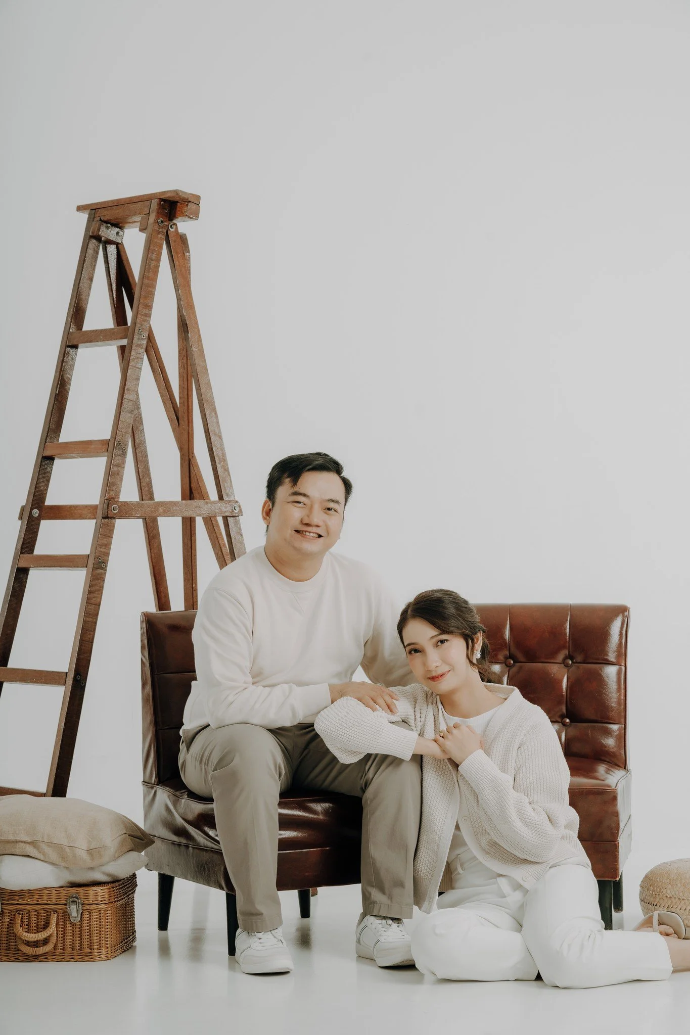 A smiling man and woman sitting on a brown leather bench in a room with a white wall and an empty wooden ladder. The woman is sitting on the floor, leaning on the man, with her hands clasped together.