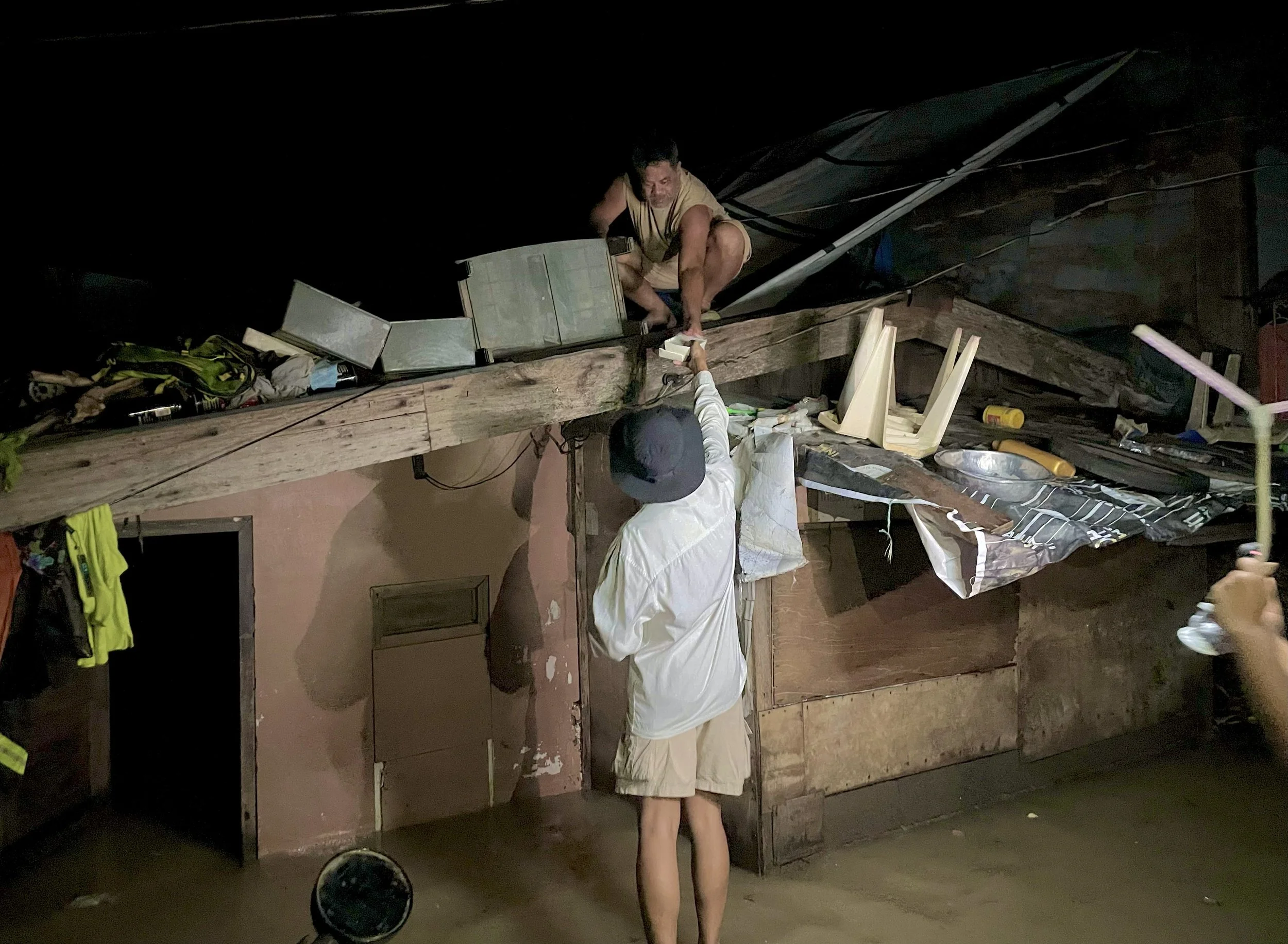 Two men working on a wooden structure, one on the roof and one on the ground, in a dimly lit environment.