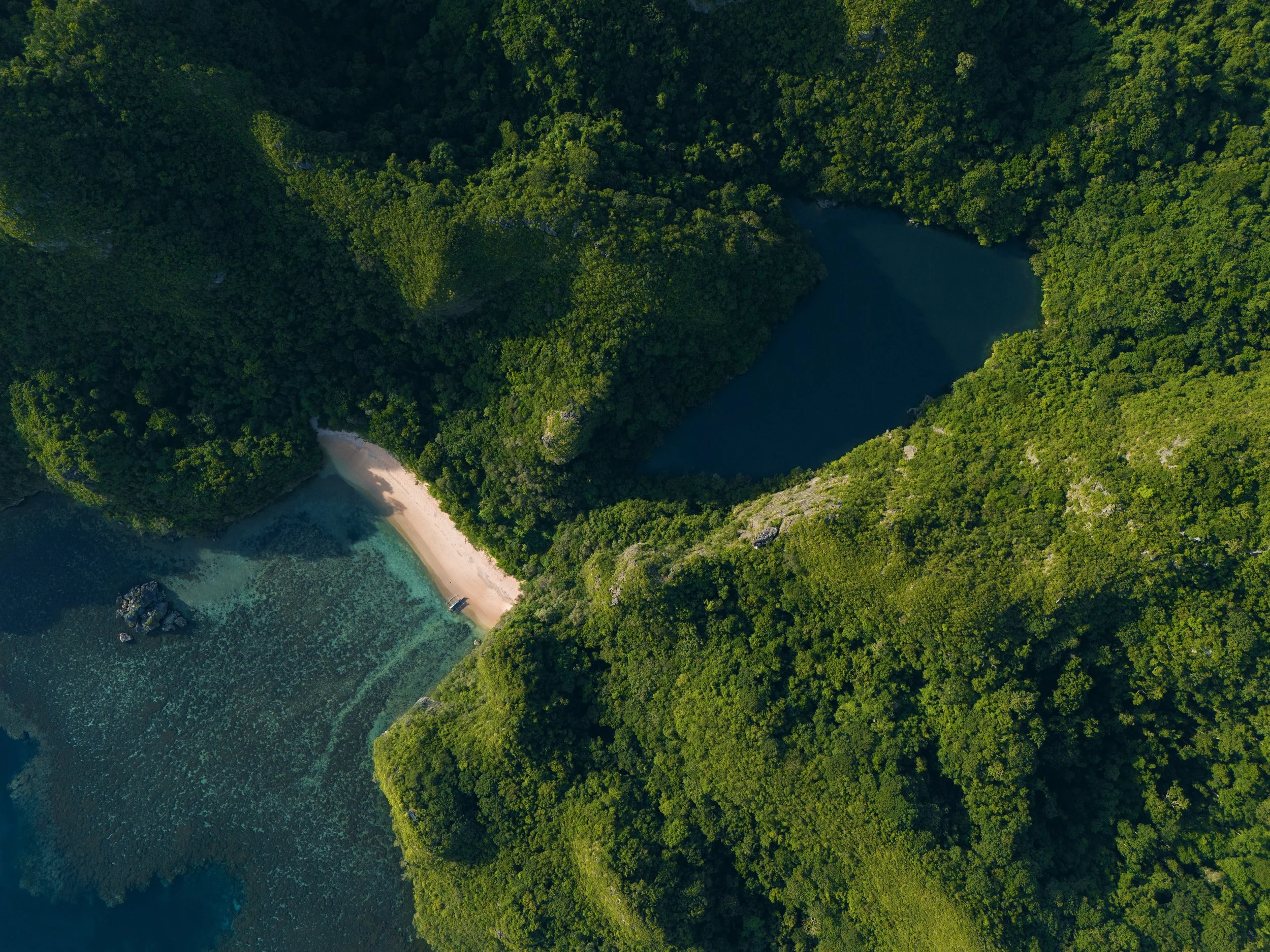 Aerial view of a lush green island with a small beach and a boat, surrounded by clear blue water and dense forested hills.