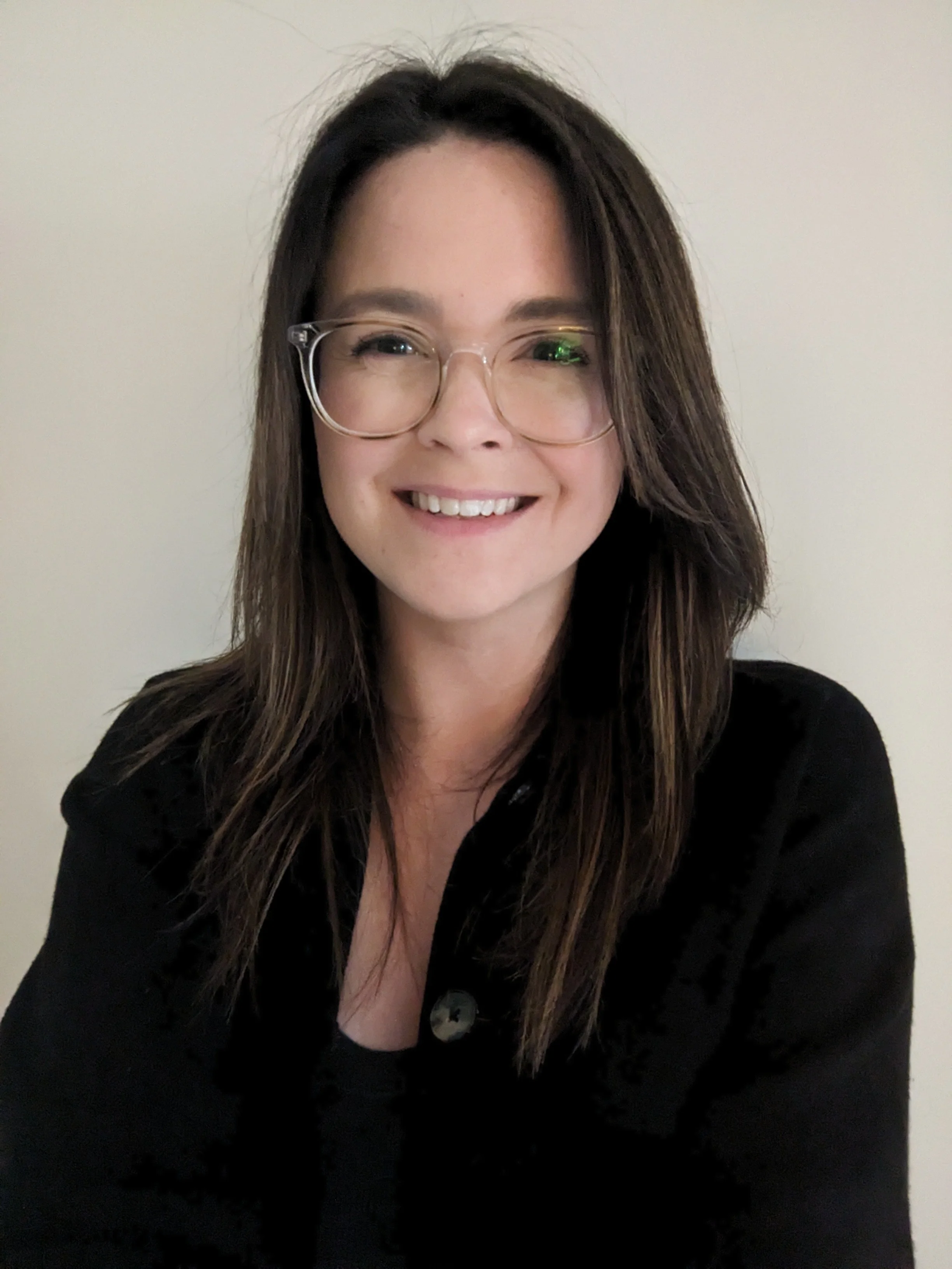 A woman with long dark hair, glasses, and a black top smiling at the camera against a plain light-colored background.