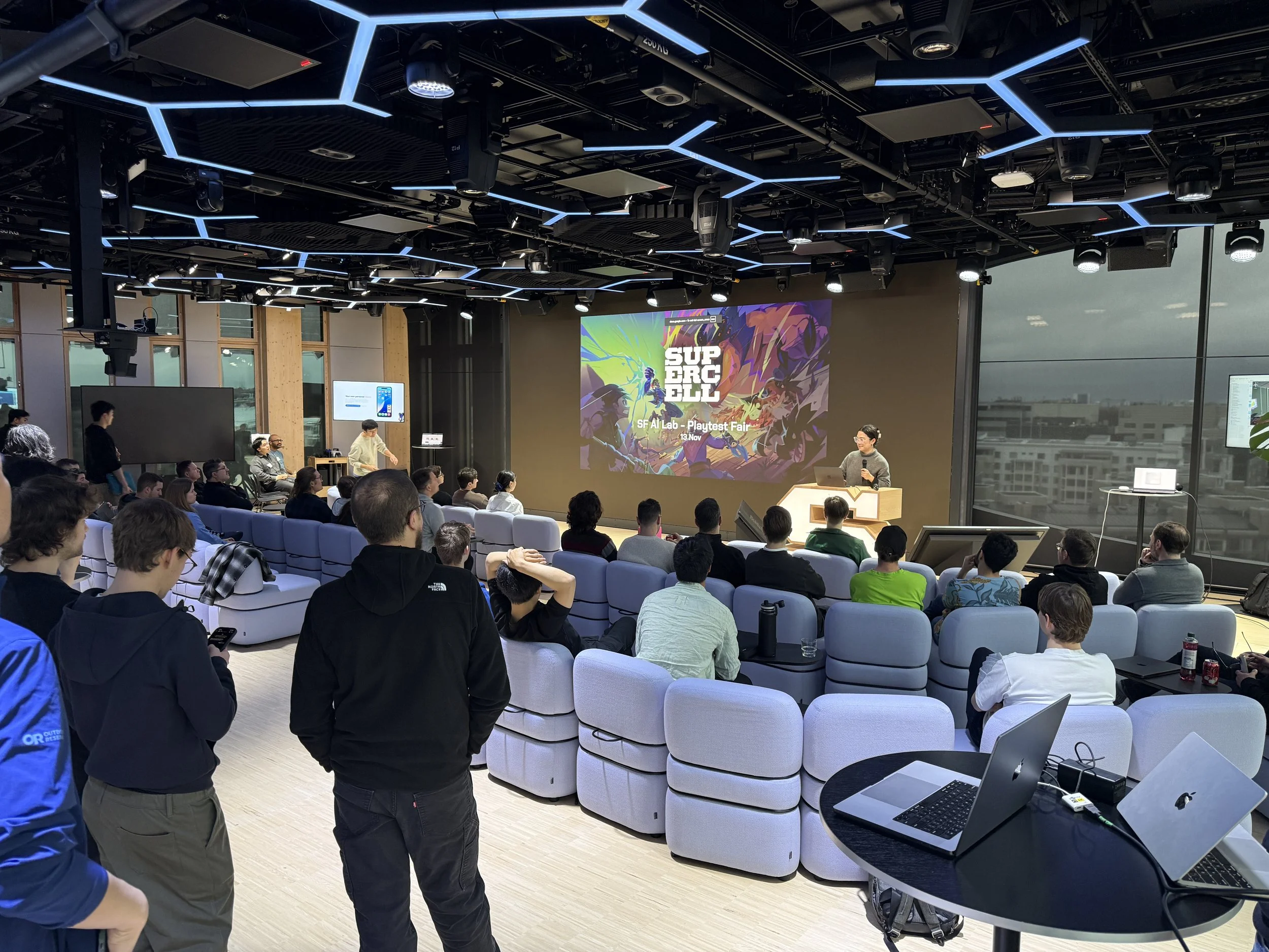 A presentation or event taking place in a modern conference room with a large screen displaying graphics for 'Supercell Playtest Fair'. There are many attendees seated, some standing, listening to the speaker at the podium. The room features large windows, a ceiling with blue LED lights, and laptops on tables at the front.