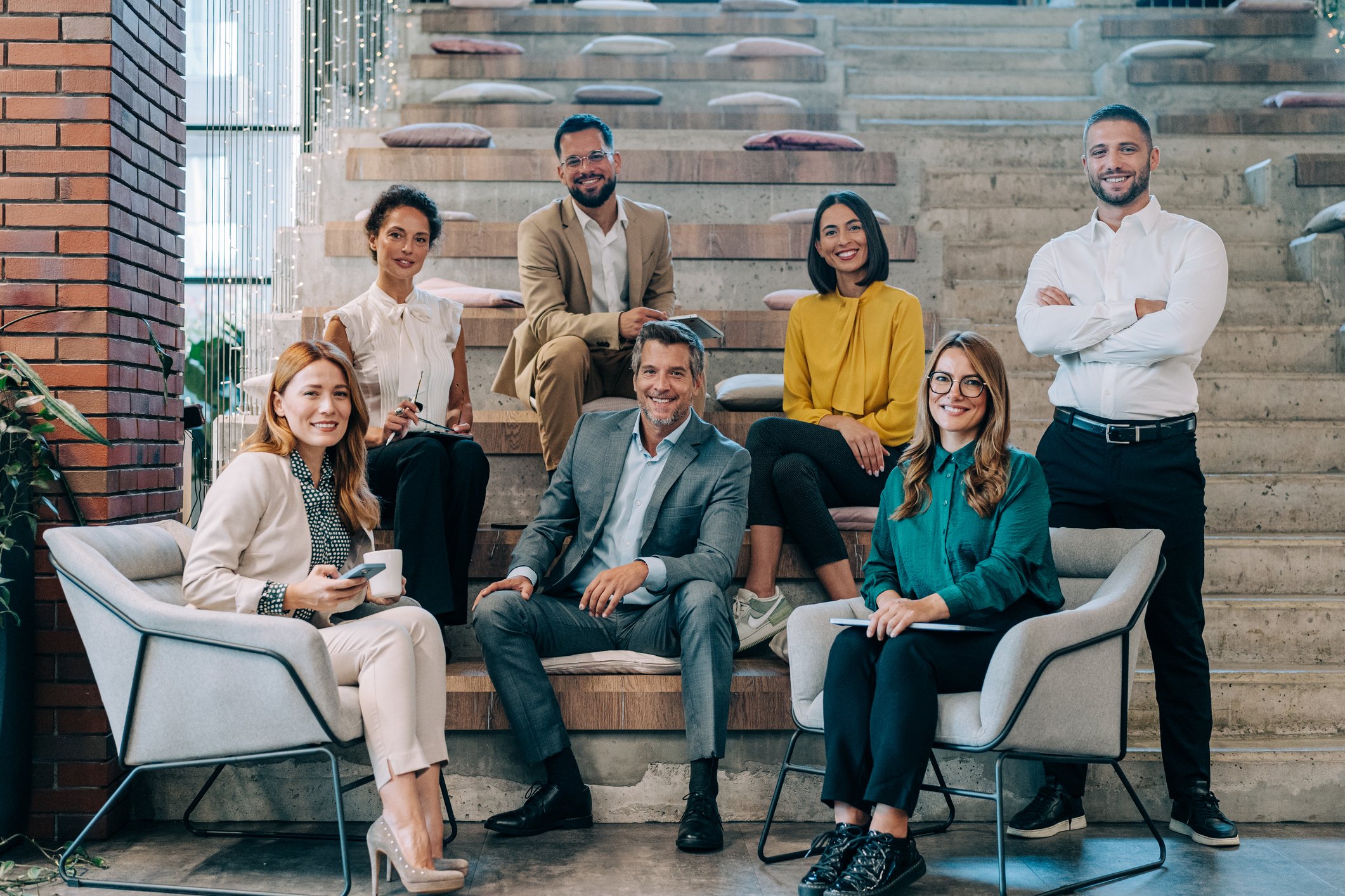 Group of seven diverse professionals posing for a photo indoors, sitting and standing on a staircase with cushions, in a modern office space.