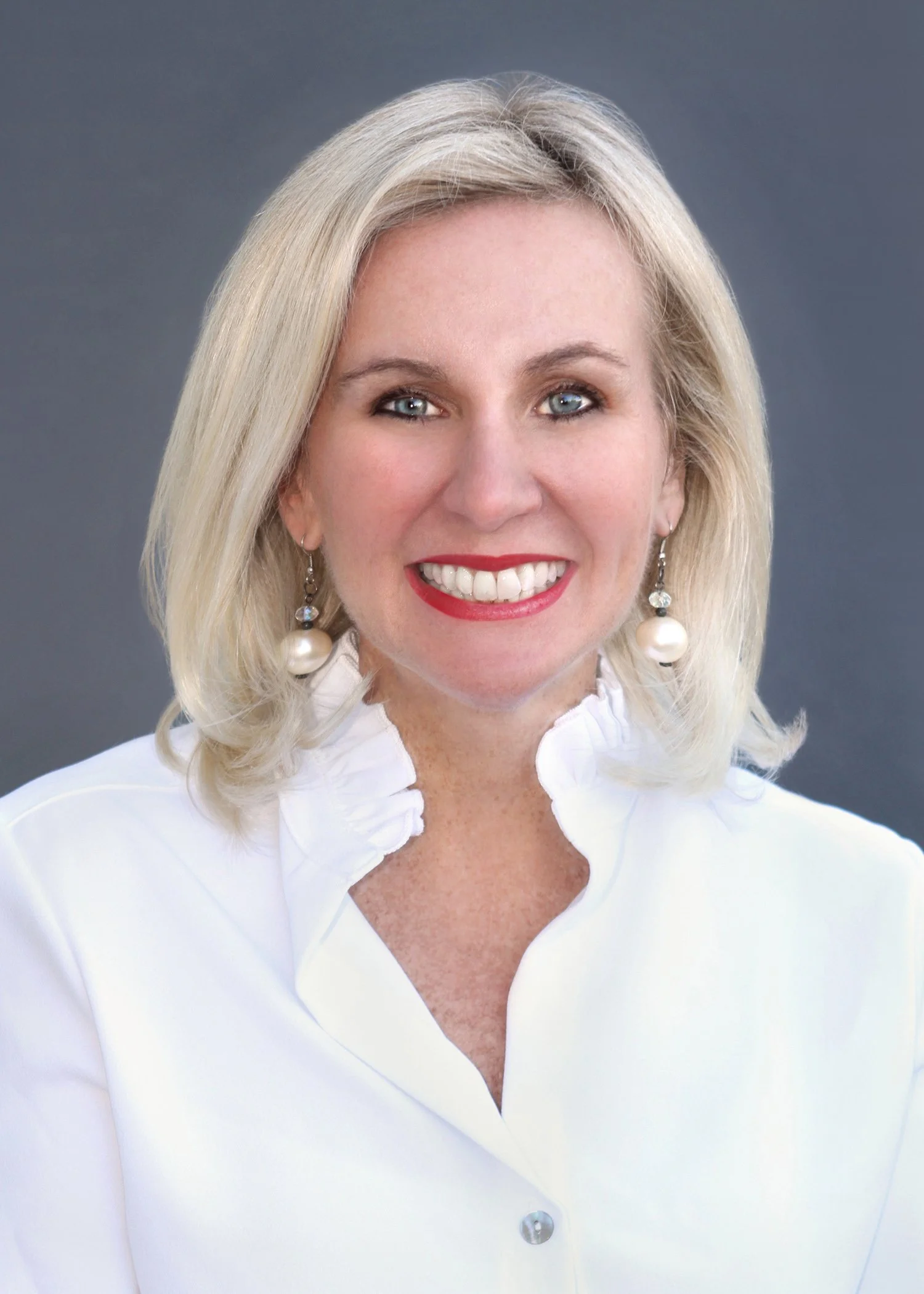 A woman with blonde hair wearing a white blouse, red lipstick, and pearl earrings, smiling against a dark background.