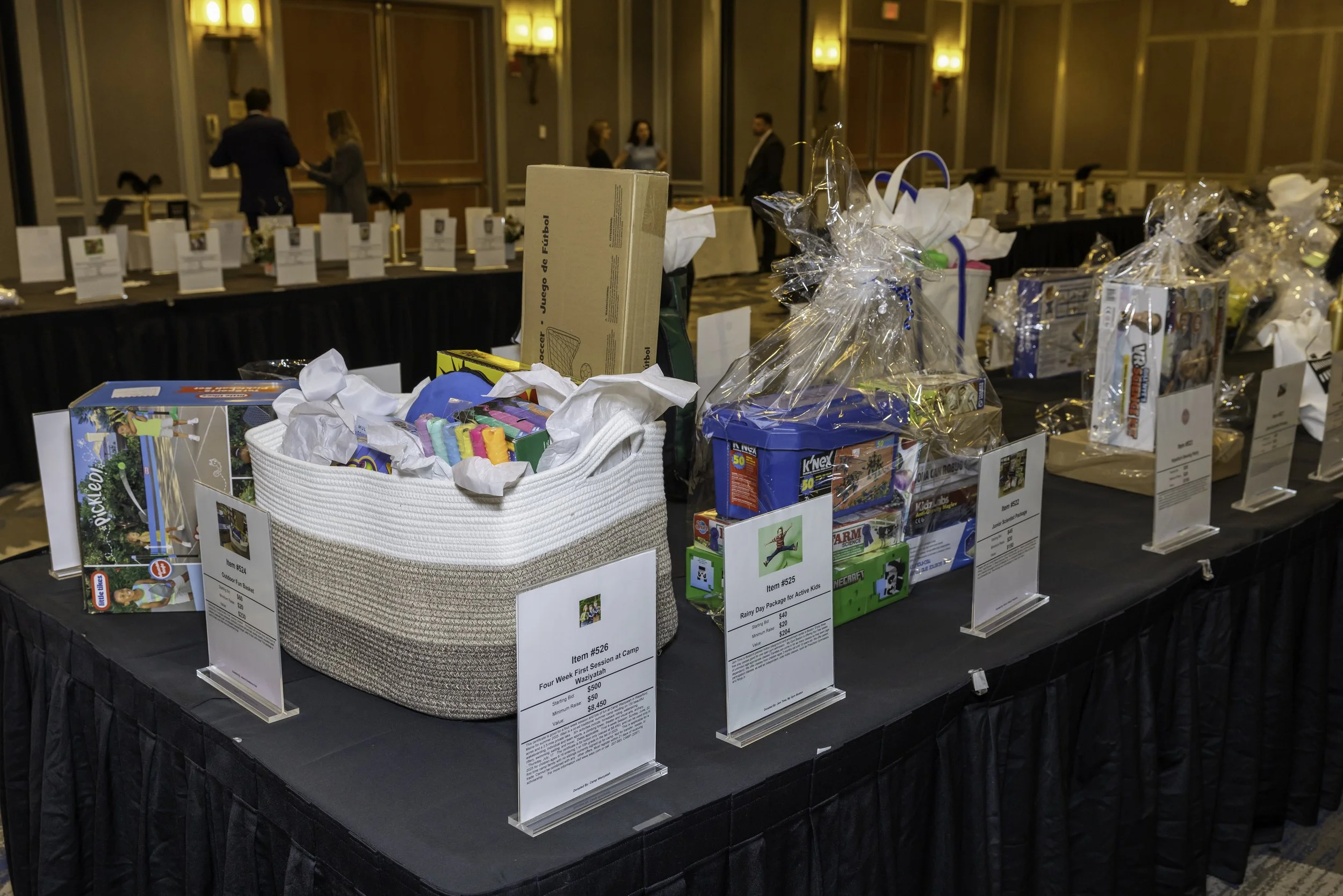 Auction display table with wrapped gift baskets and boxed toys in a hotel conference room.