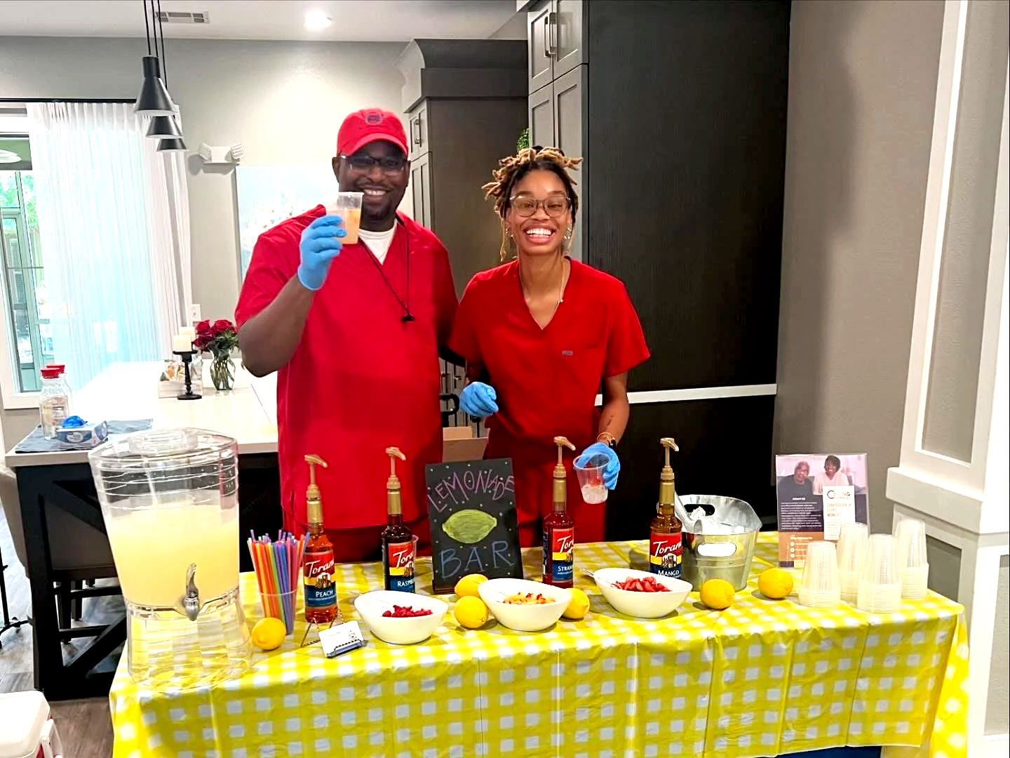 Two smiling people standing behind a lemonade stand wearing red shirts and gloves, with a large pitcher of lemonade, lemons, bowls of strawberries, and bottles of Torani syrup on a yellow checkered tablecloth, inside a bright kitchen.