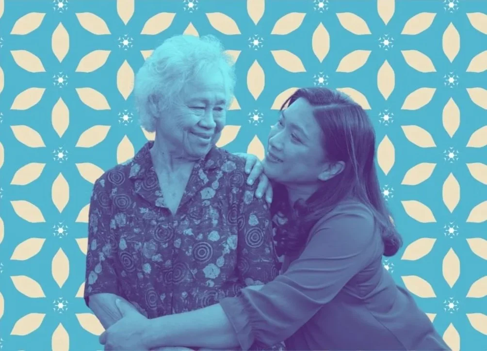 Two women, one elderly with white hair and one younger with dark hair, smiling and embracing each other in front of a decorative backdrop with a geometric pattern and snowflake motifs.