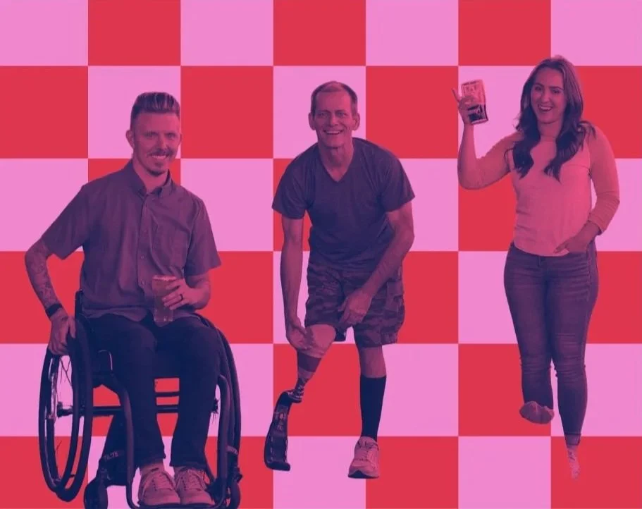 Three people smiling and posing against a pink and red checkered background. One person is in a wheelchair, while the others are standing, with one holding a drink.
