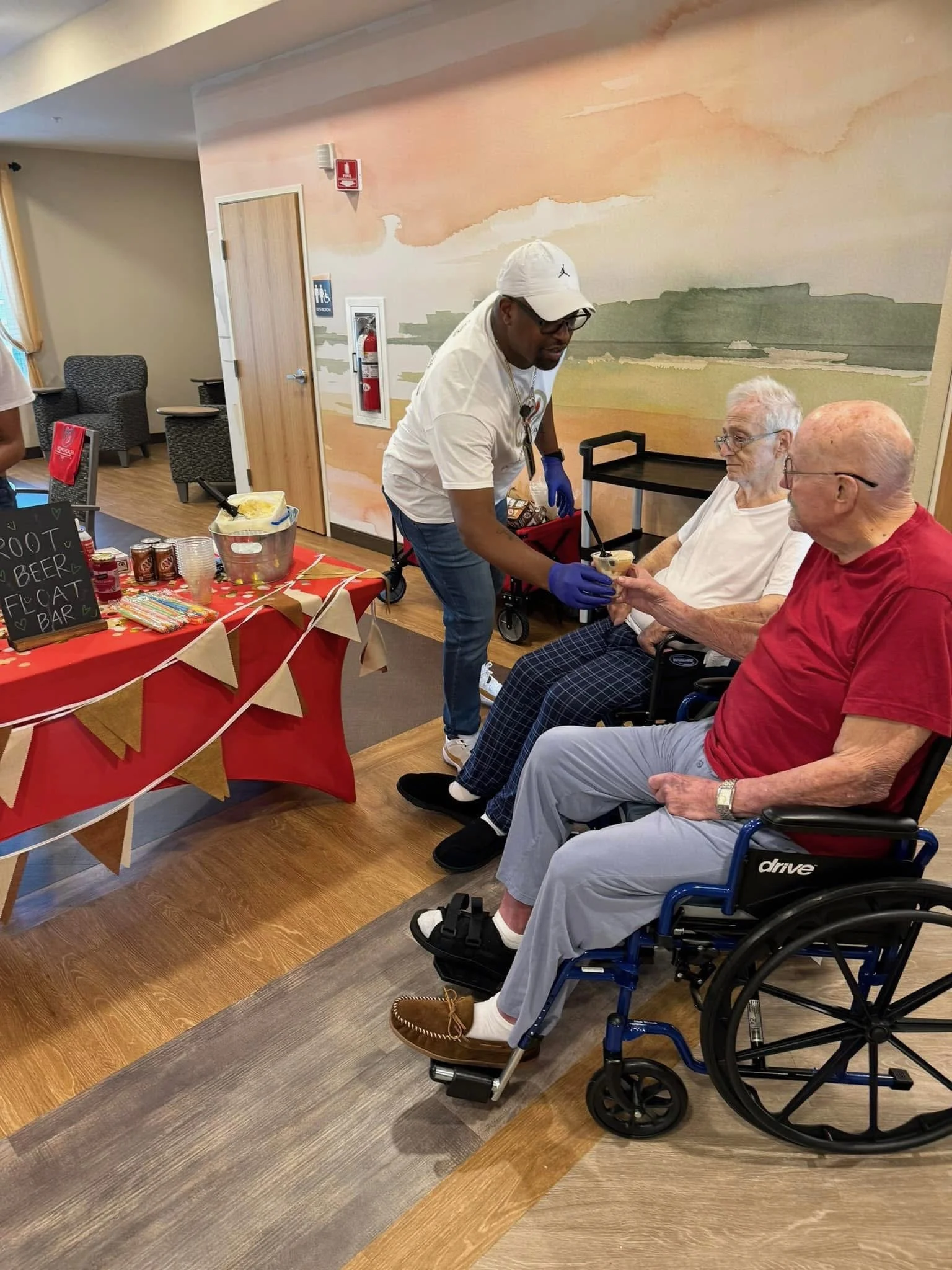 A caregiver in a white shirt and cap hands a dessert to an elderly man in a wheelchair, with another elderly woman sitting beside him, at a community event with a decorated table in the background.