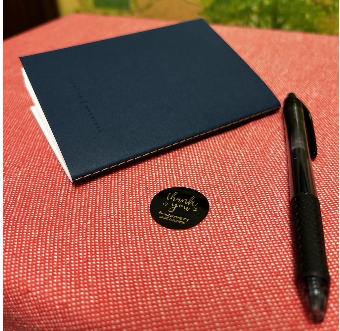 A blue notebook, a black pen with textured grip, and a round black sticker with gold text thanking for supporting a small business, placed on a pink textured surface.