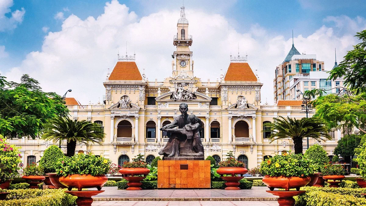Historical building with a clock tower, ornate architecture, and statues, surrounded by greenery and a bronze statue of a seated woman reading a book in front.