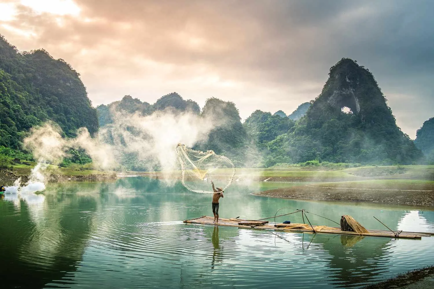 A person fishing on a bamboo raft on a river surrounded by green mountains with mist in the air.
