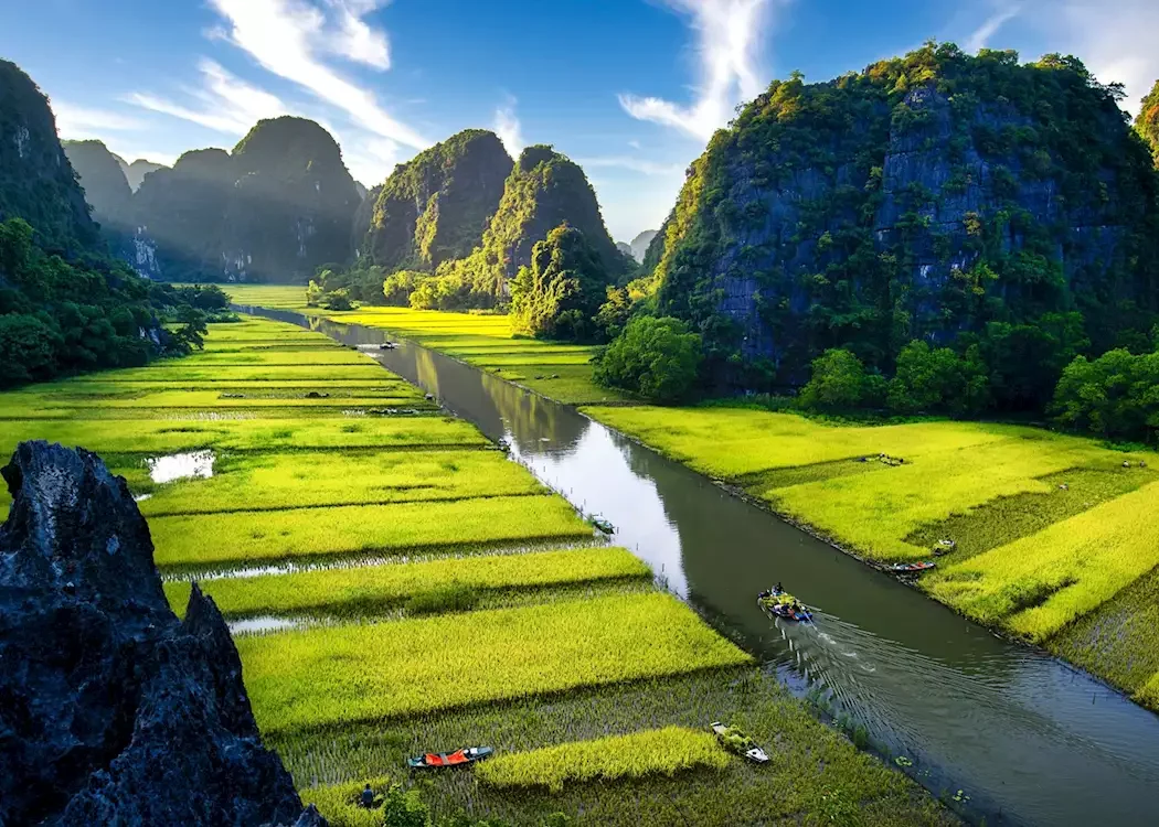 Lush green rice paddies with boats on a river, surrounded by tall, green-studded limestone karsts under a partly cloudy sky.