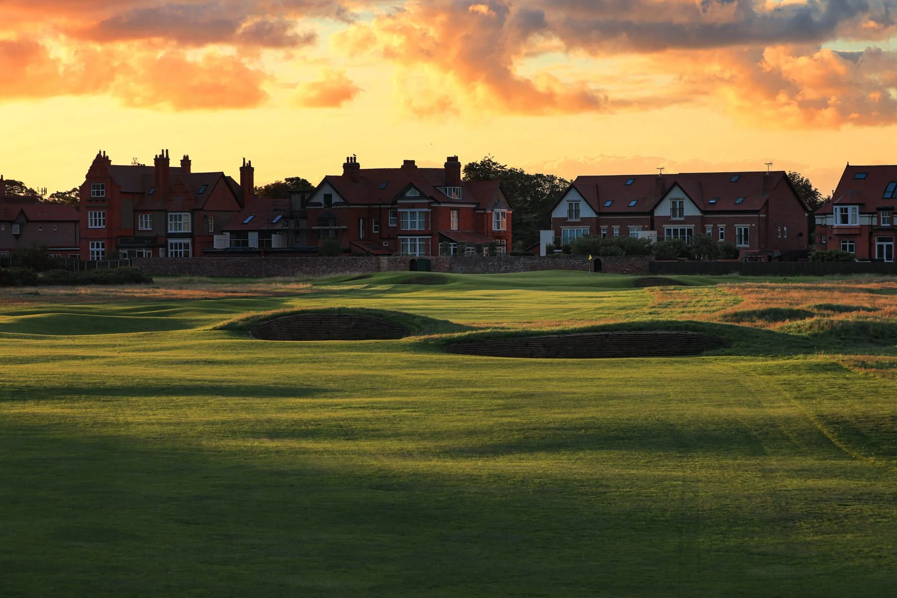 A scenic view of a golf course at sunset with green fairways, sand bunkers, and a row of large brick houses with chimneys and gabled roofs in the background under a partly cloudy sky.