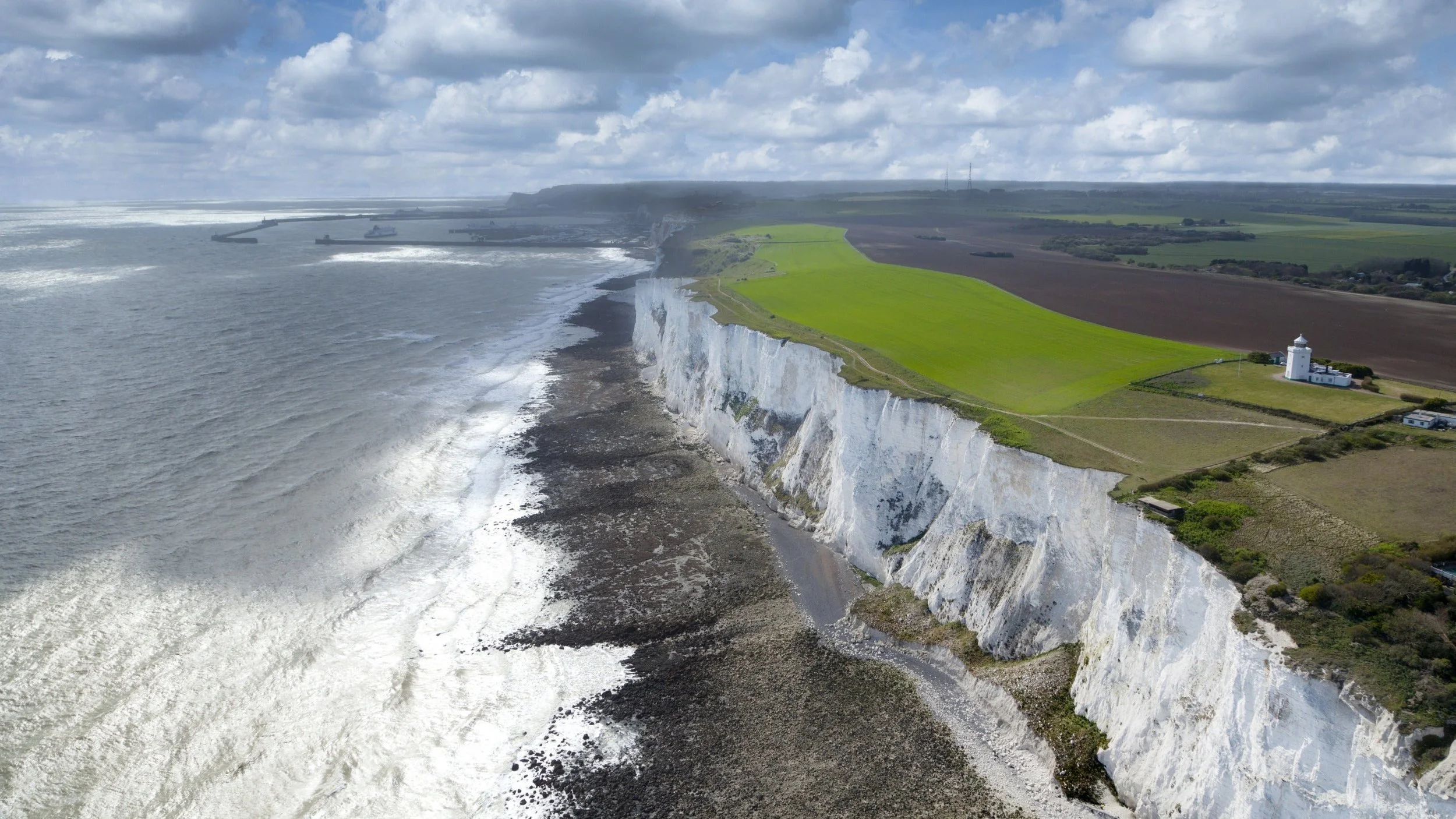 An aerial view of white chalk cliffs along the coastline, with a lighthouse near the edge, green fields behind, and ships out at sea under a partly cloudy sky.