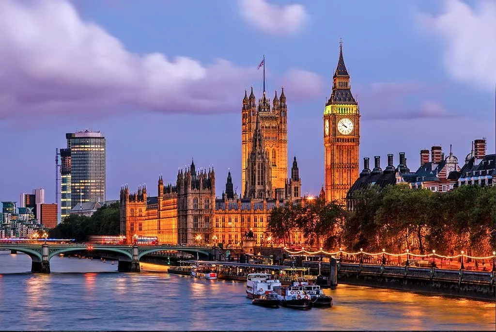 Twilight view of the Palace of Westminster, also known as the Houses of Parliament, in London, England, with Big Ben clock tower, river Thames in foreground, boats, and illuminated walkway.