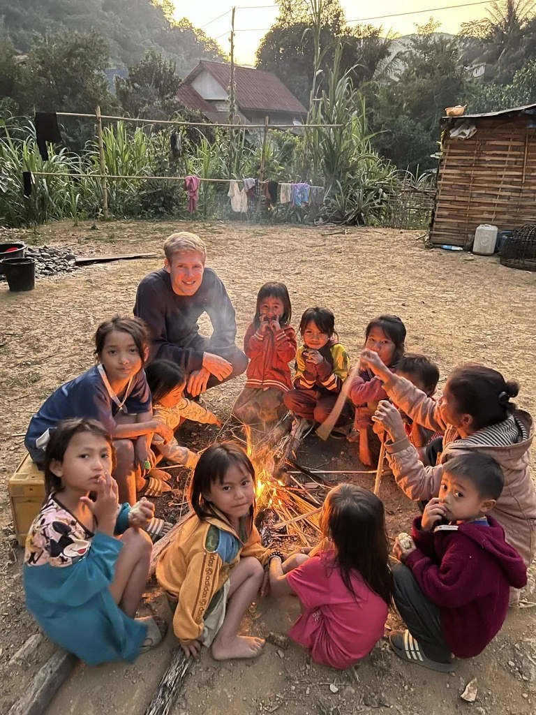 Eine Gruppe von Kindern und einem Erwachsenen sitzen im Kreis um ein kleines Feuer im Freien. Es ist Abend, und im Hintergrund sind Häuser, Vegetation und Wäscheleinen zu sehen.