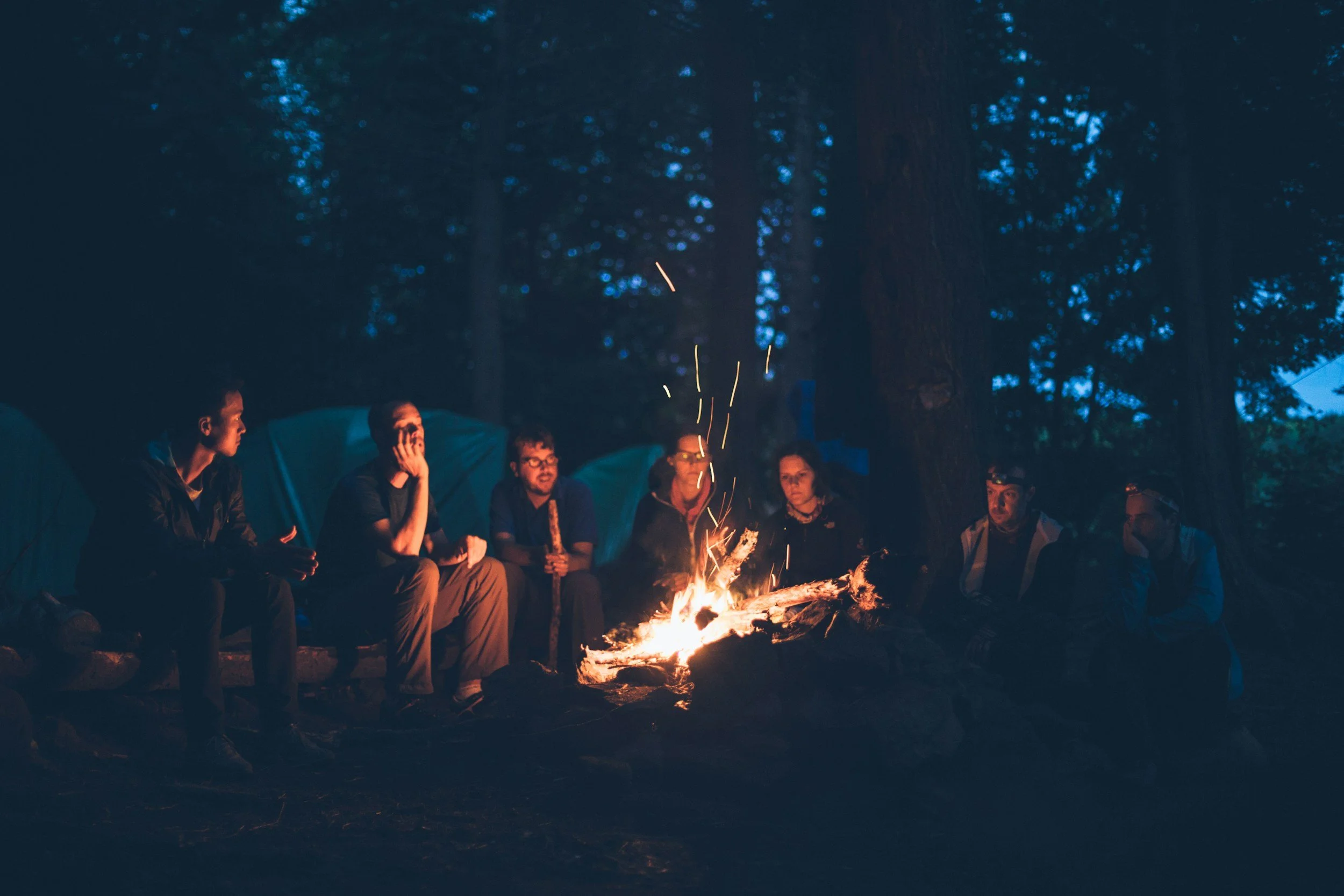 Gruppe von Menschen sitzt um ein Lagerfeuer im Freien bei Nacht, umgeben von Bäumen.