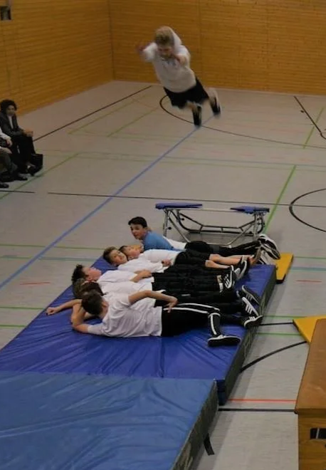 Trainer springt in einer Turnhalle auf eine Weichmatte, während mehrere Jungen darauf liegen.