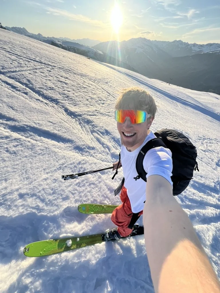 Gründer mit Ski-Ausrüstung auf Schneefeld in den Bergen, Sonnenuntergang im Hintergrund, trägt eine weiße T-Shirt, rote Shorts, reflektierende Skibrille und Rucksack, macht ein Selfie.