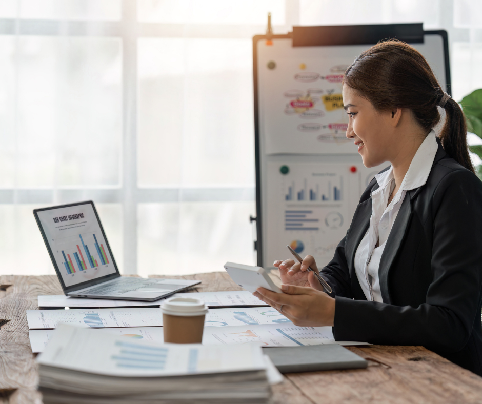 Businesswoman working at her desk with a laptop displaying bar graphs, papers, a coffee cup, and a whiteboard with charts in the background.