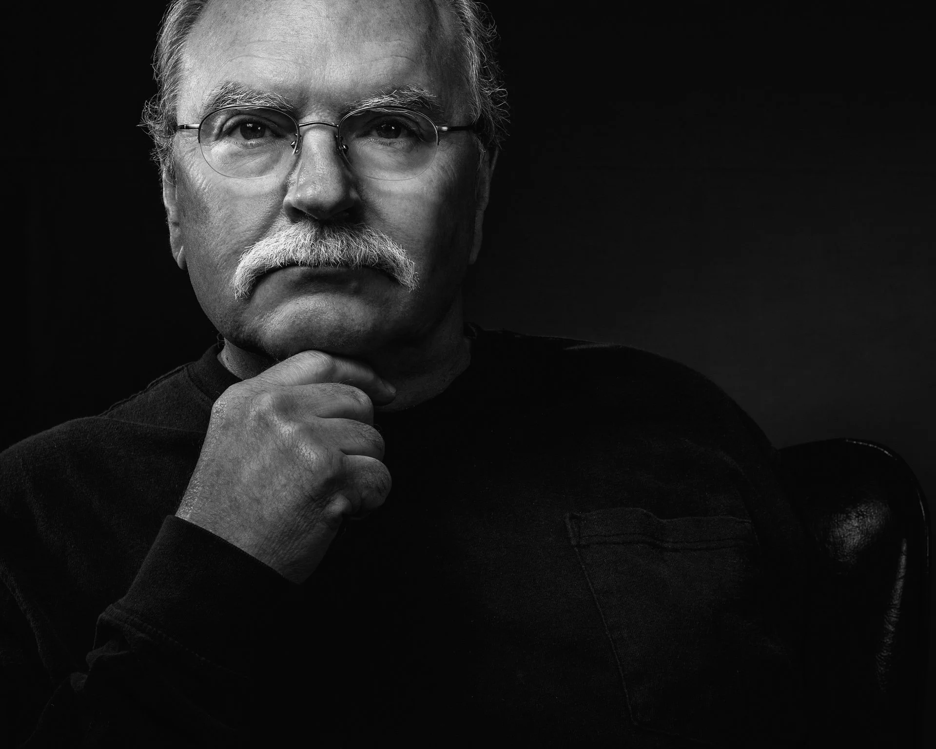 Black and white portrait of an elderly man with glasses and a mustache, looking thoughtfully into the camera, with his hand resting on his chin, wearing a dark shirt.