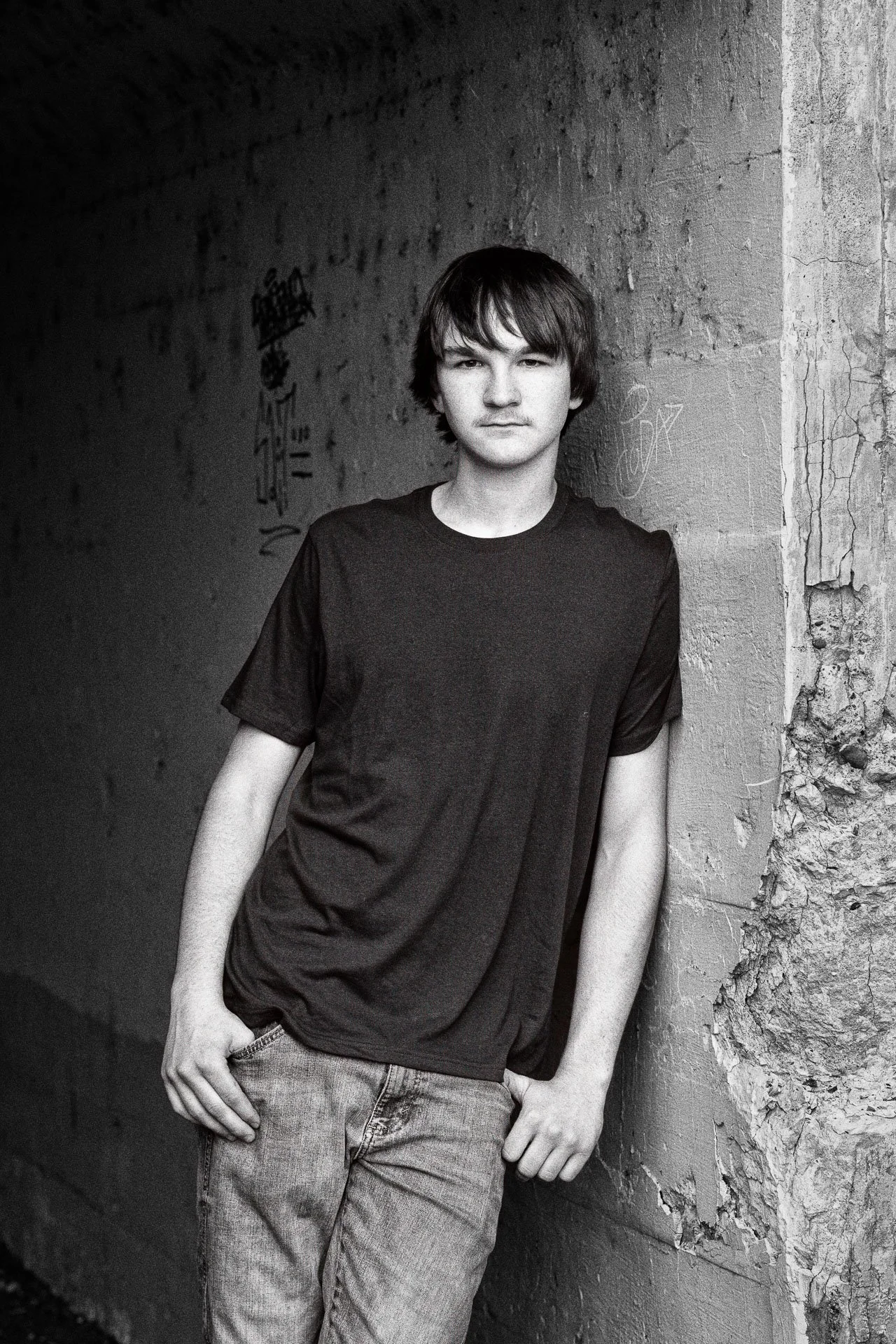 A young man with dark hair and light skin leaning against a textured wall in an alley, looking at the camera with a neutral expression, wearing a black T-shirt and jeans, in black and white.