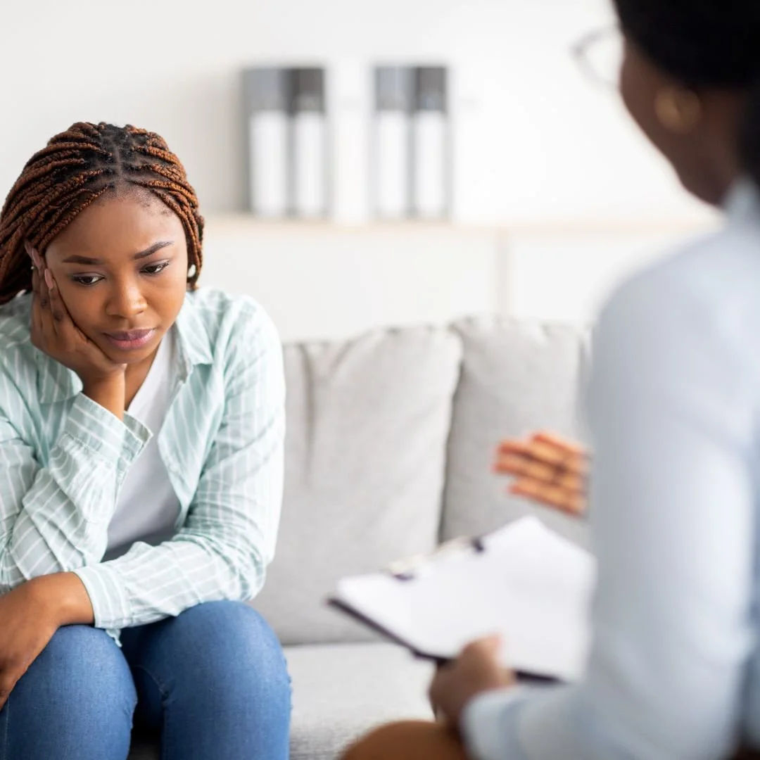 A young woman sitting on a couch with her hand on her face, looking worried while talking to a therapist or counselor who is holding a clipboard.