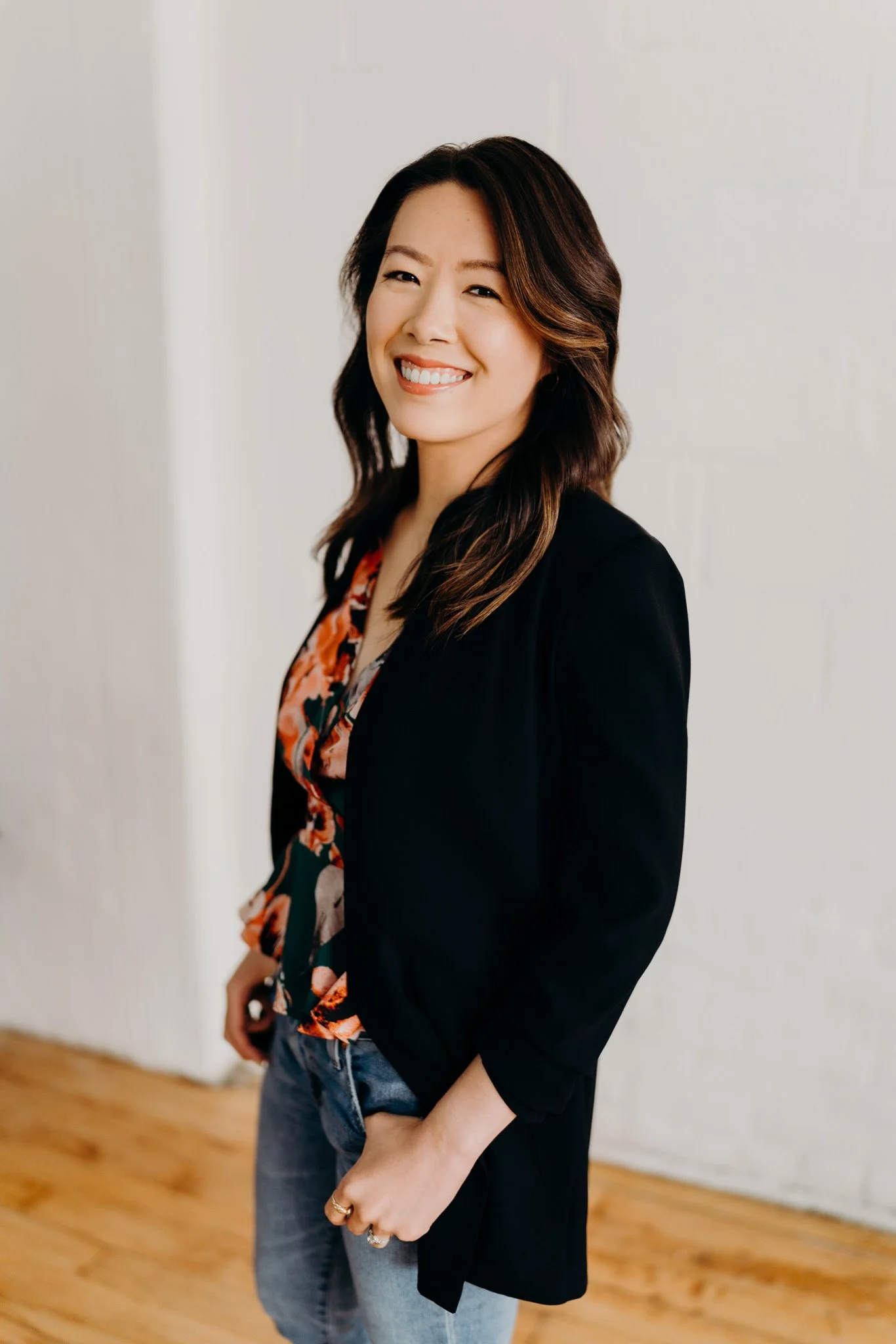 Dr. Kathy Zhang: A smiling Asian woman with shoulder-length dark hair with highlights, wearing a black blazer over a floral blouse and jeans, standing in front of a white wall and light wooden floor.