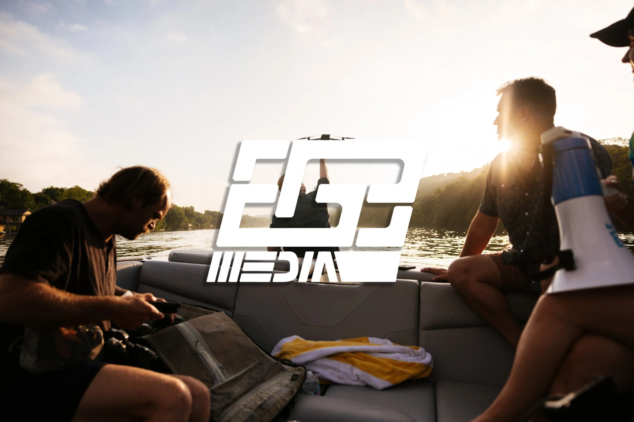 People sitting on a boat near a lake during sunset, with a man looking at his phone, and others sitting and chatting, water and trees in the background.