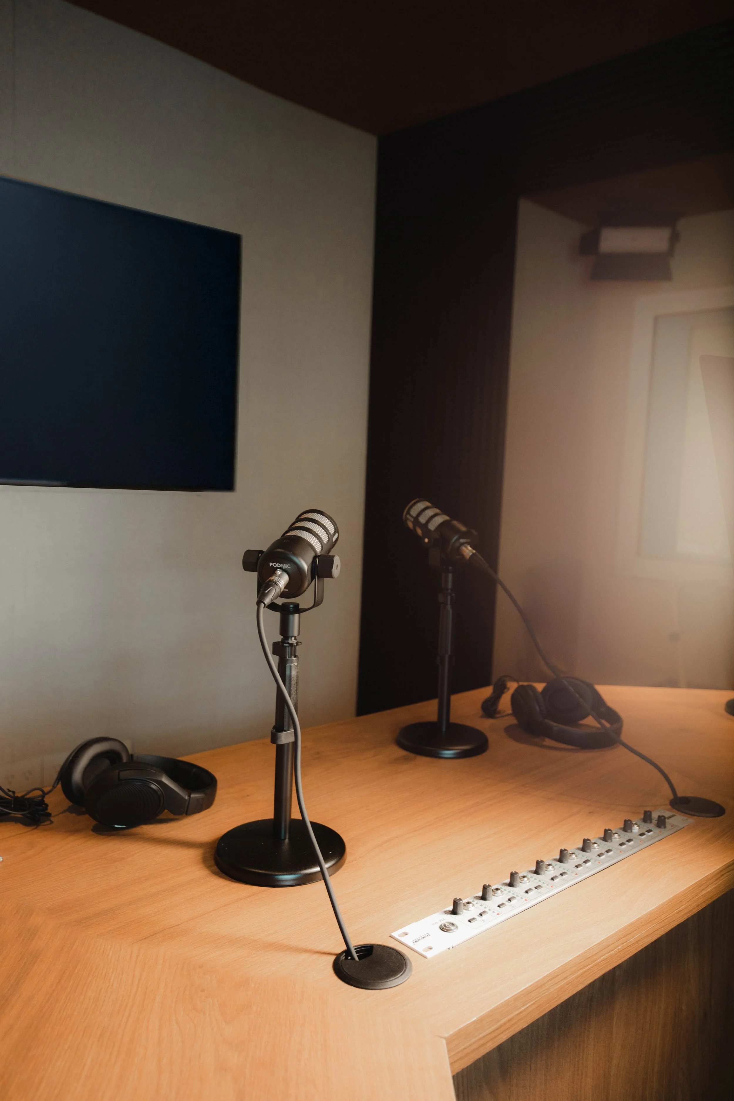 Estudio de grabación con micrófonos, audífonos y mesa de mezclas sobre mesa de madera, en un espacio con paredes en tonos neutros y pantalla en la pared.