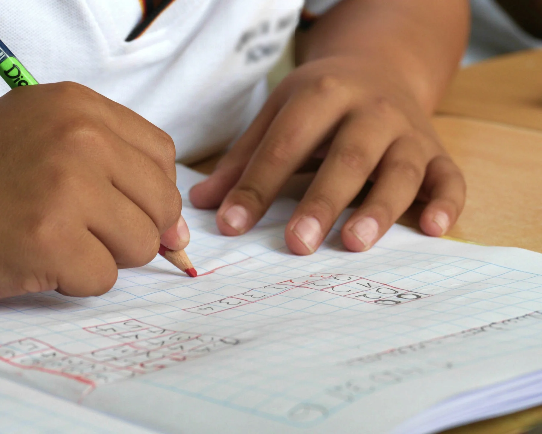A child's hand is drawing and writing on graph paper with a red colored pencil, with the child's other hand resting on the paper.