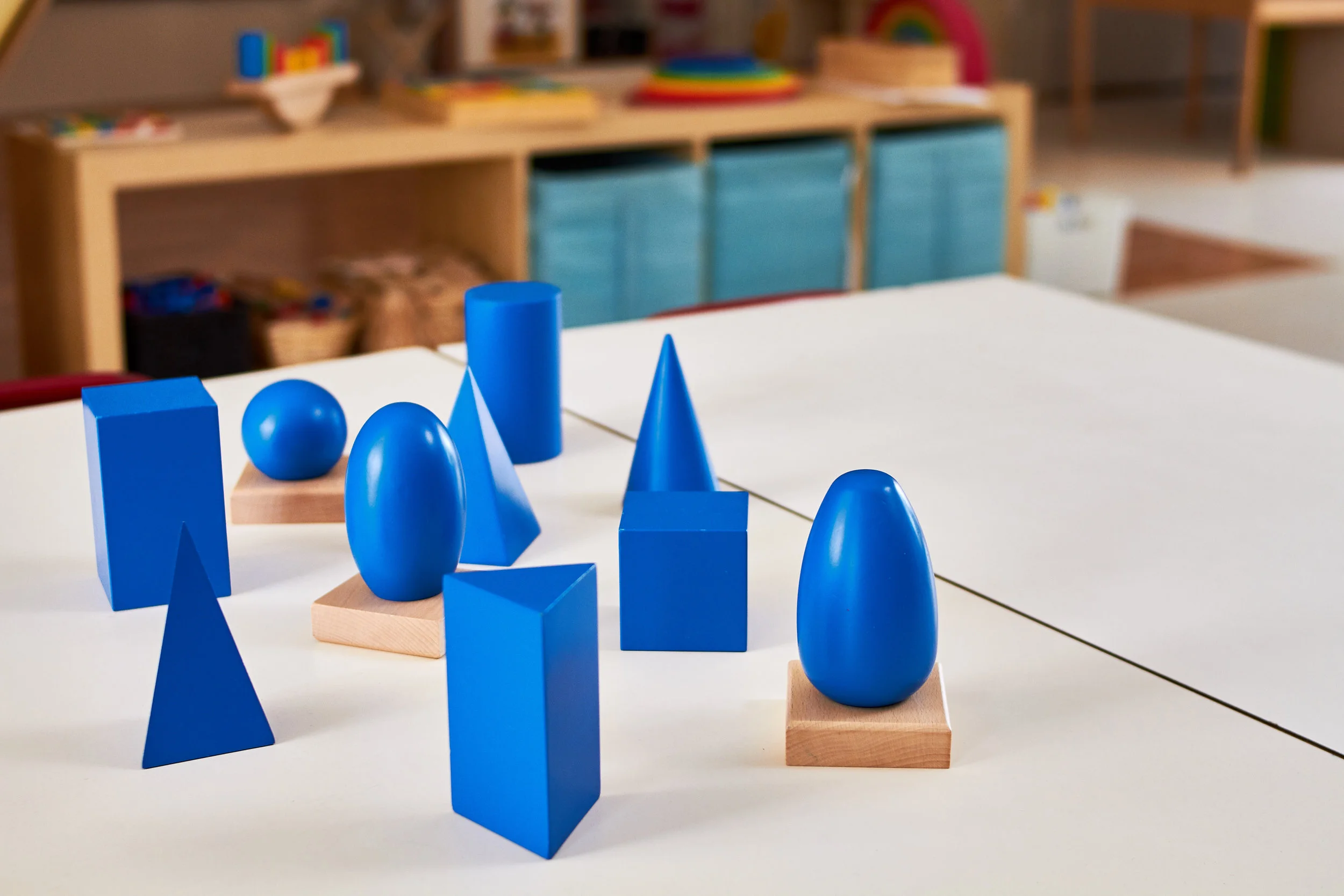 Blue geometric wooden shapes, including spheres, cylinders, cones, and rectangular blocks, arranged on a white table in a classroom setting with shelves in the background.