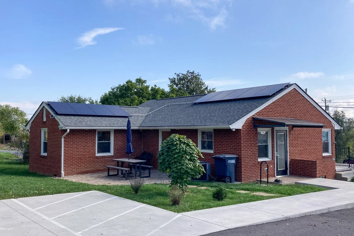A brick house with solar panels on the roof, a small outdoor patio with a picnic table and umbrella, green grass, a tree, and trash cans in front.