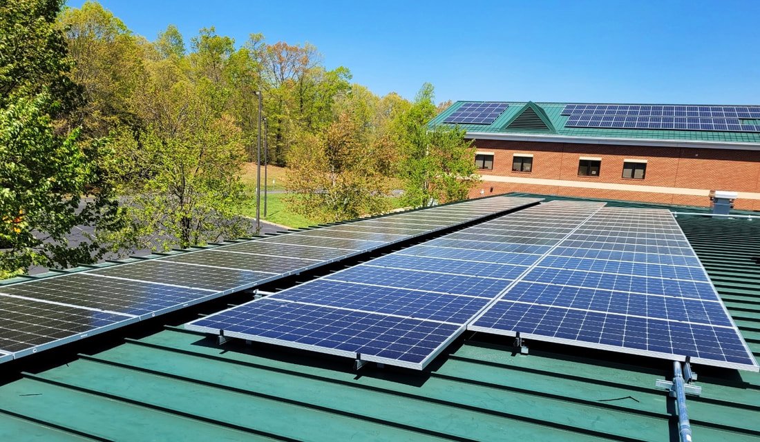 Multiple solar panels installed on a green roof with a background of trees and a brick building with solar panels on its roof.