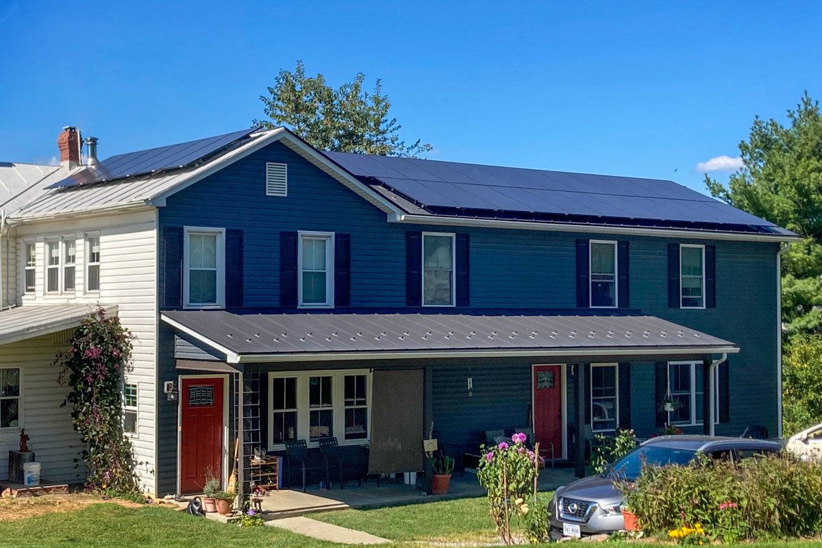 A two-story house with blue and white siding, a metal roof with solar panels, and a small front porch with chairs and potted plants. There are cars parked in the driveway and garden with flowers in front.