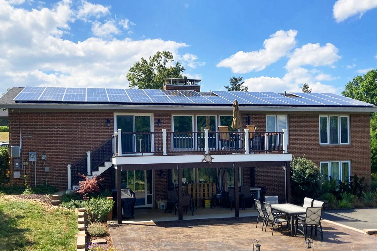 A two-story brick house with solar panels on the roof, outdoor patio furniture, and a landscaped yard under a partly cloudy sky.