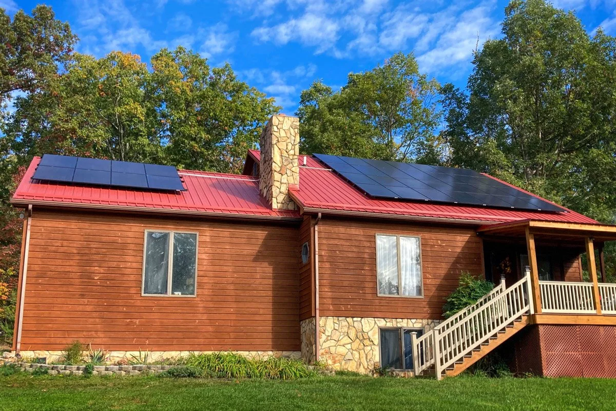 A two-story house with a red metal roof and solar panels. The house has wooden siding, a stone chimney, and a front porch with stairs. There are two windows visible on the front, and the house is surrounded by green grass and trees with autumn foliag