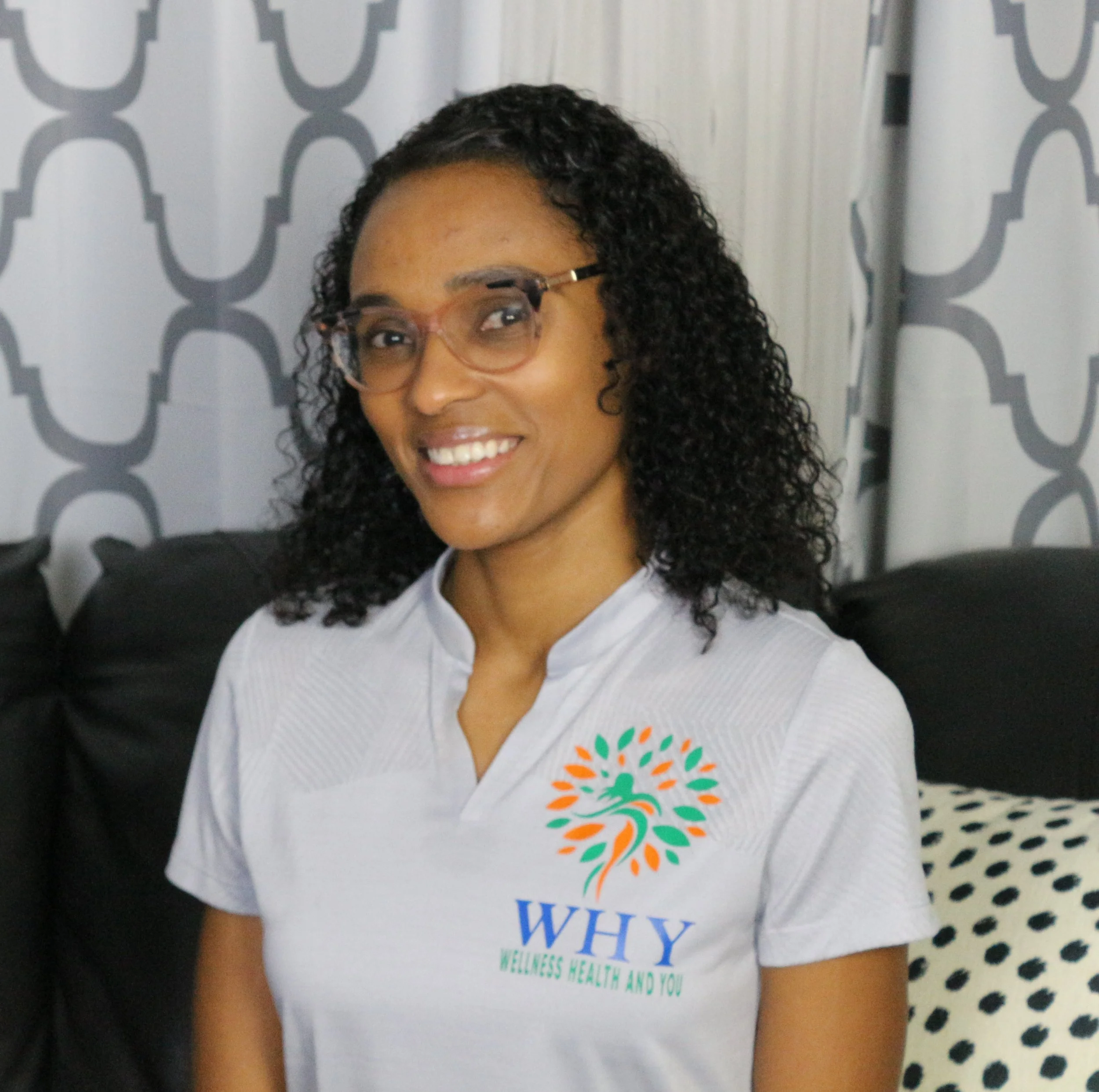 Smiling woman wearing a white polo shirt with a colorful logo that reads 'WHY Wellness Health Advocates,' sitting on a black couch with patterned curtains in the background.