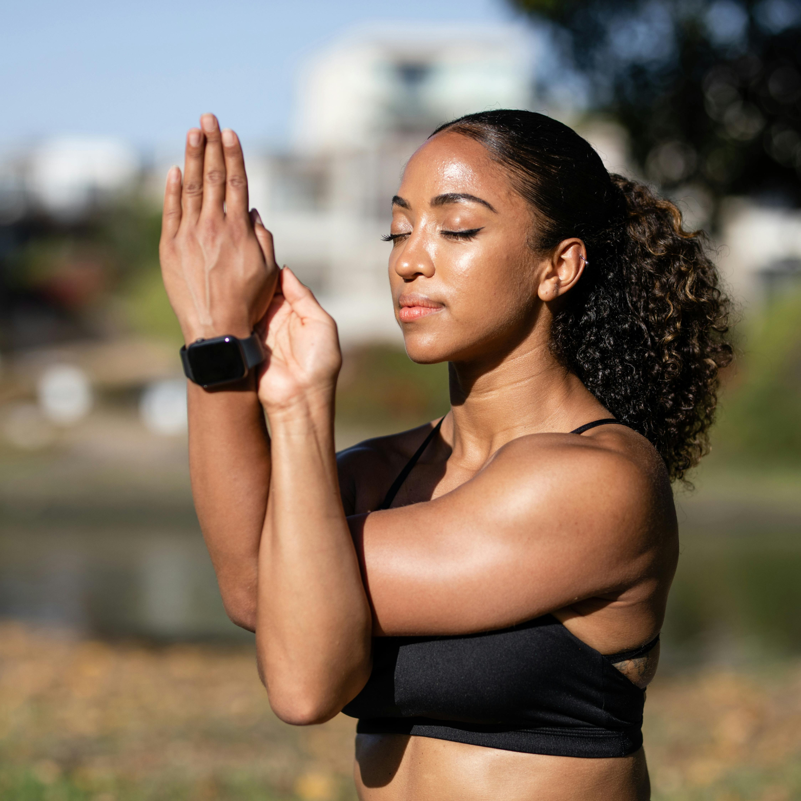Young woman with curly hair in a black sports bra doing yoga outdoors in bright sunlight, with her eyes closed and hands in prayer position.