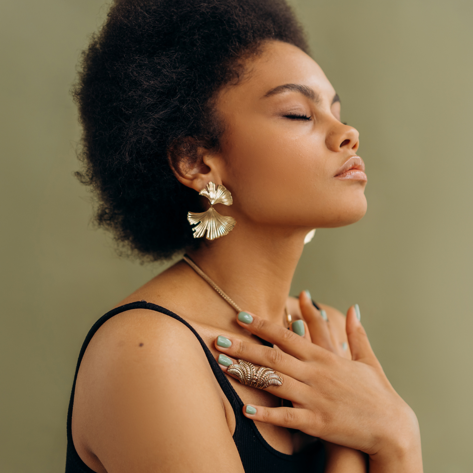 A woman with natural curly hair wearing statement gold earrings, a ring, a necklace, and painted fingernails, with her eyes closed and hand on her chest against a muted green background.