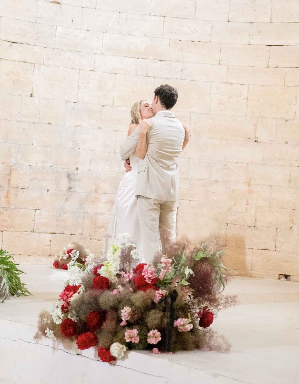 A bride and groom share a kiss on their wedding day, standing on a platform decorated with a variety of red, pink, white, and purple flowers in front of a beige brick wall.