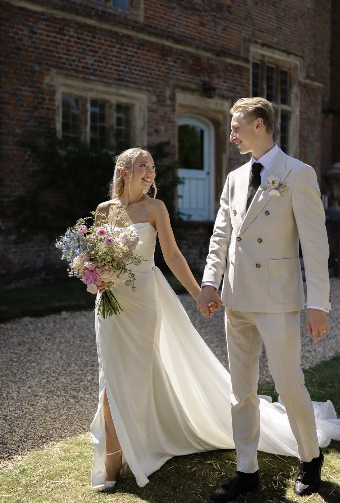 A bride and groom holding hands outdoors on their wedding day, smiling at each other, with a brick building in the background. The bride wears a white strapless wedding gown and holds a bouquet of pink, purple, and white flowers. The groom wears a white suit with a black tie and a boutonniere.