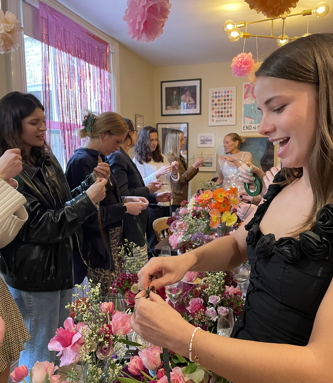 A group of women gathered in a room with pink and orange flowers, decorating or arranging flowers on a table. The room is decorated with pink tissue paper pom-poms, hanging lights, and framed pictures on the wall.