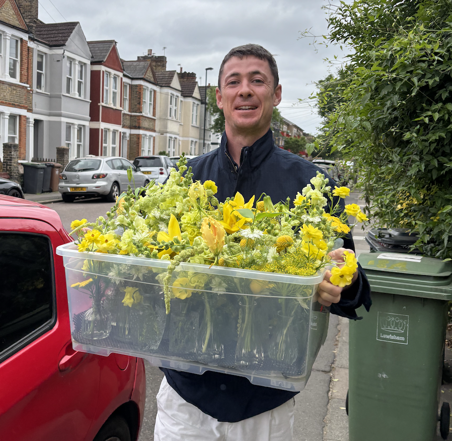 A man smiling and holding a clear plastic container filled with yellow flowers on a residential street with parked cars and houses.